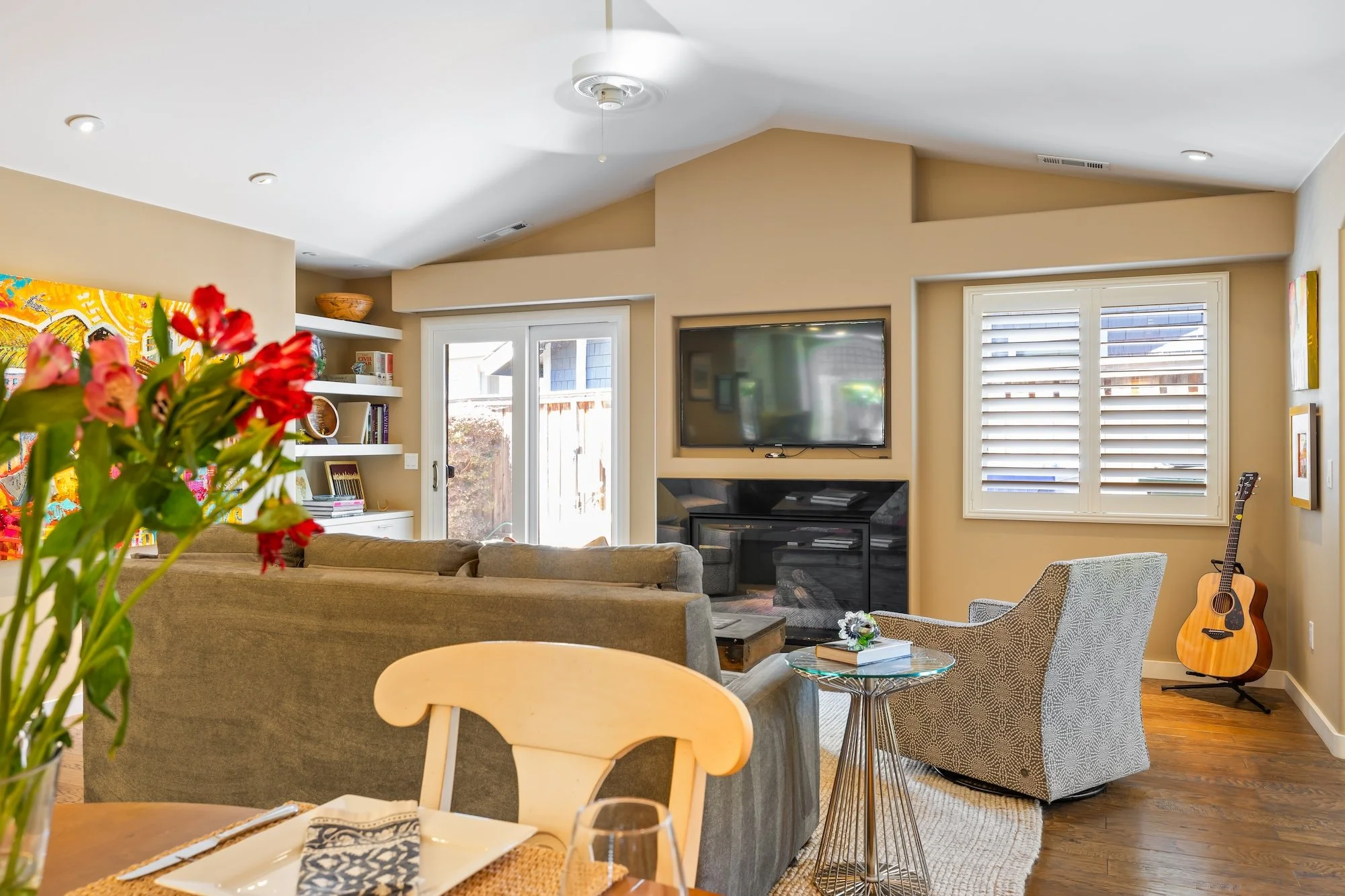 A cozy living room with a beige sofa, a patterned gray armchair, a guitar, a flat-screen TV above a fireplace, and a sliding glass door letting in natural light. There are also bookshelves, colorful artwork, and decorative items.