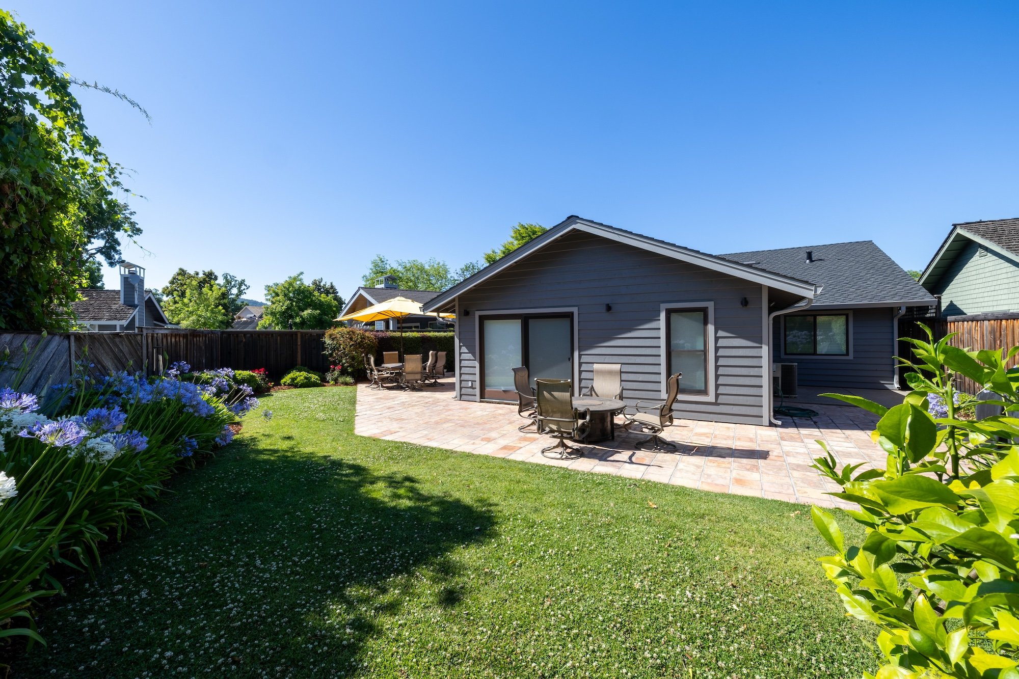 A backyard patio with outdoor dining tables, chairs, and an umbrella, surrounded by a green lawn, colorful flowers, and neighboring houses under a clear blue sky.