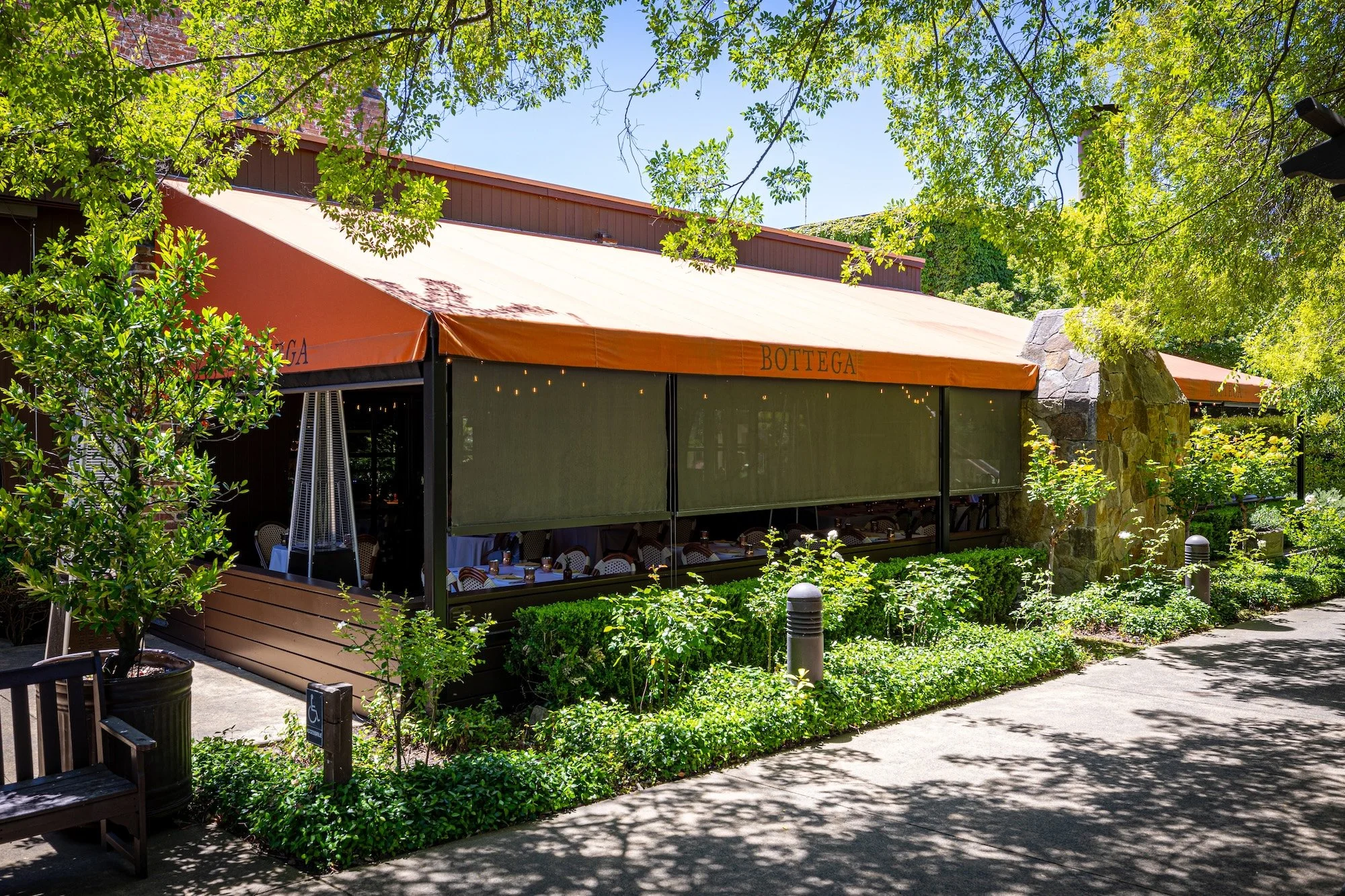Outdoor restaurant patio with tables set for dining, surrounded by lush green trees and bushes, with a bright orange canopy that has the word 'BOTTEGA' written on it. There is a stone structure, possibly a fireplace, to the right side.