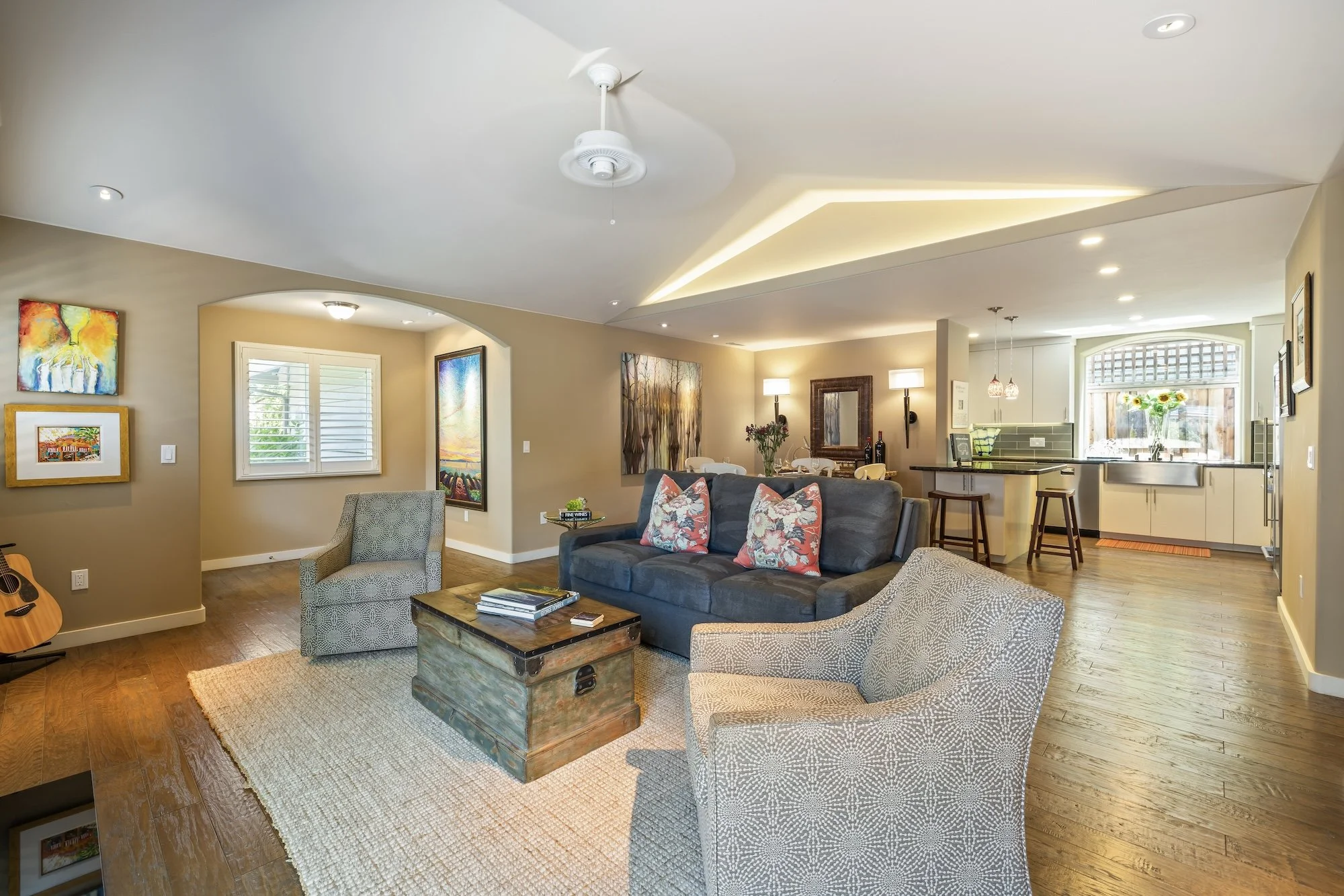 Living room with seating area, colorful paintings on beige walls, hardwood floor, kitchen visible in the background, ceiling fans, recessed lighting, and decorative pillows on a dark sofa.