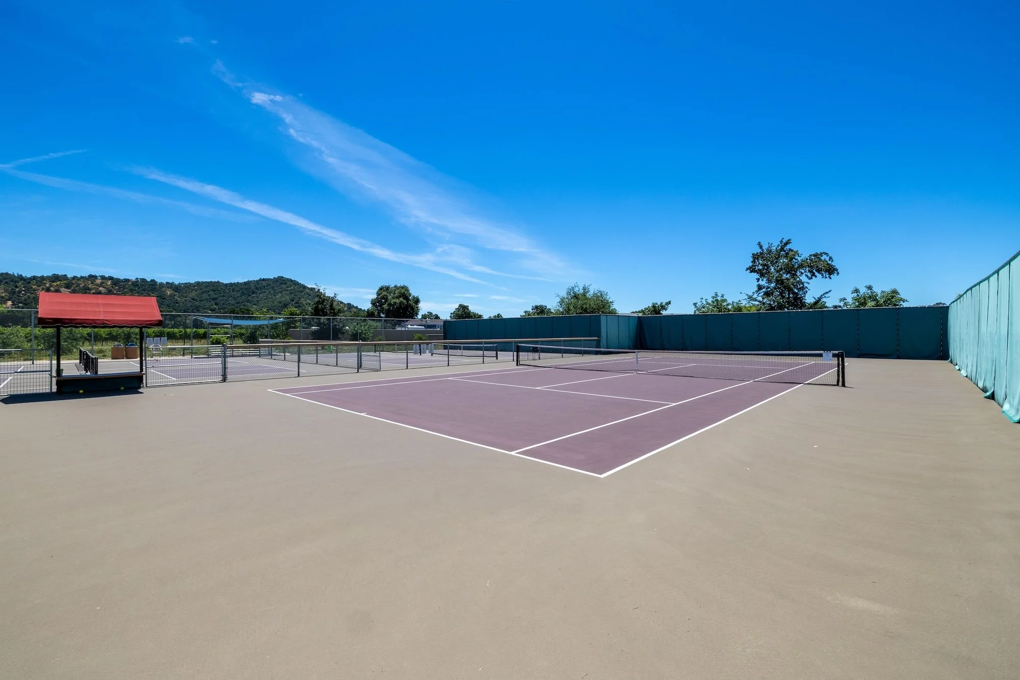 An empty outdoor tennis court with a cloudy blue sky and hills in the background, featuring a small red pavilion, tennis nets, and surrounding fencing.