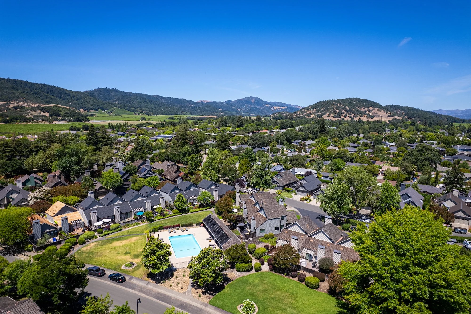 Aerial view of a suburban neighborhood with houses, a swimming pool, and lush green trees, set in a mountainous landscape under a clear blue sky.
