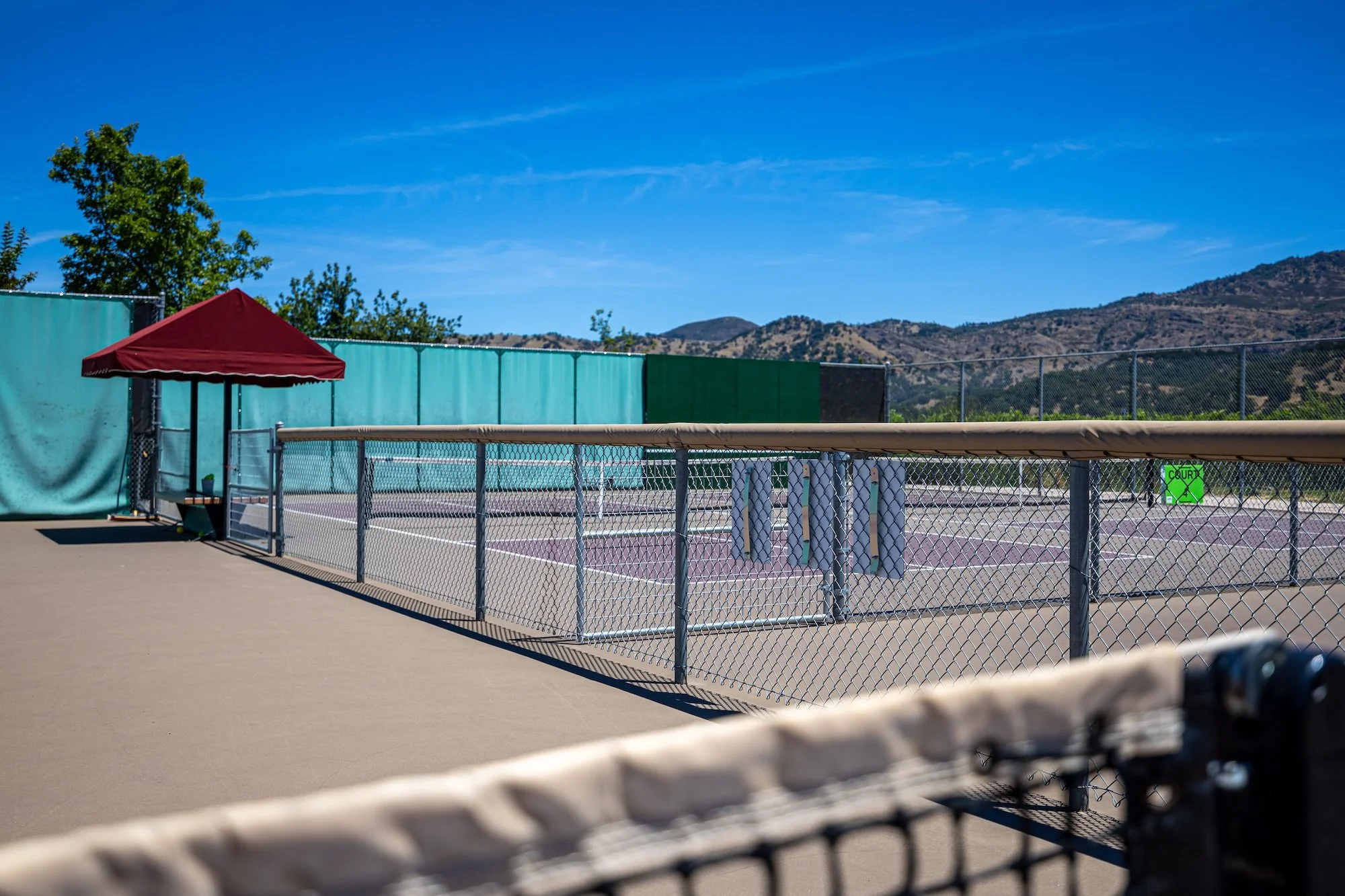 Empty tennis court with privacy screens, a red canopy, and a mountainous landscape in the background under a clear blue sky.
