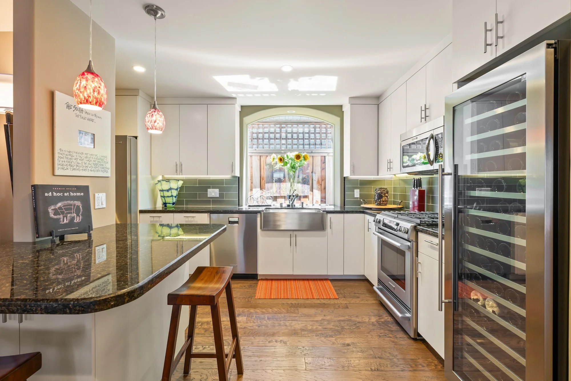 Modern kitchen with white cabinets, stainless steel appliances, green tile backsplash, and a window with sunflower flowers. There are two red pendant lights hanging from the ceiling and a granite countertop with a book and a wooden stool.