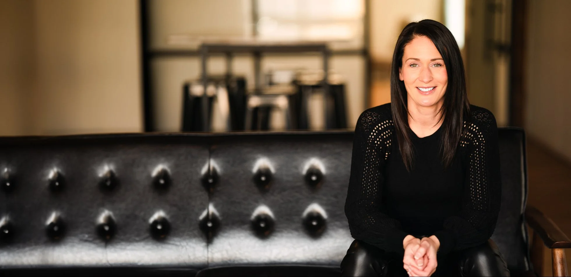 Kate sitting on a black leather tufted sofa, smiling. The background shows a blurred interior with chairs and a table.