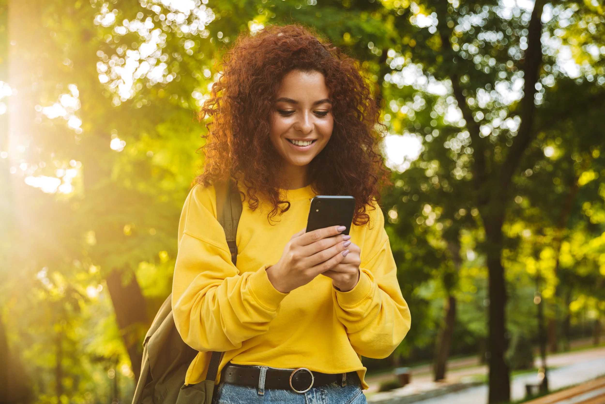 Smiling woman in yellow sweater using smartphone outdoors with trees in background