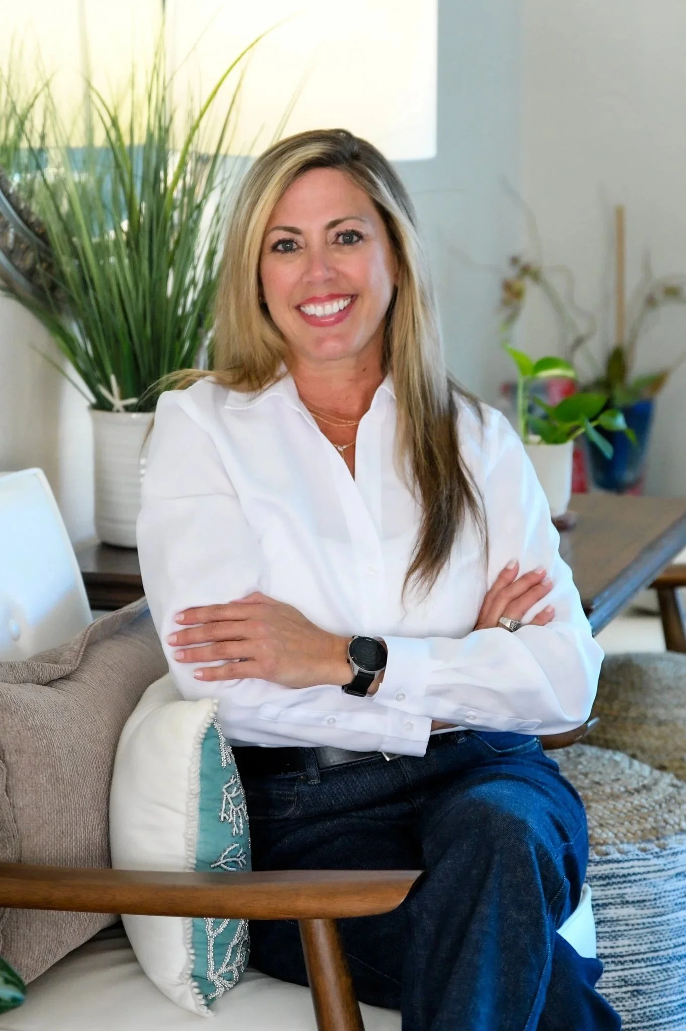 A woman with blonde hair sitting on a beige sofa, smiling with arms crossed, wearing a white shirt, dark jeans, and a black watch, with potted plants and decorative items in the background.