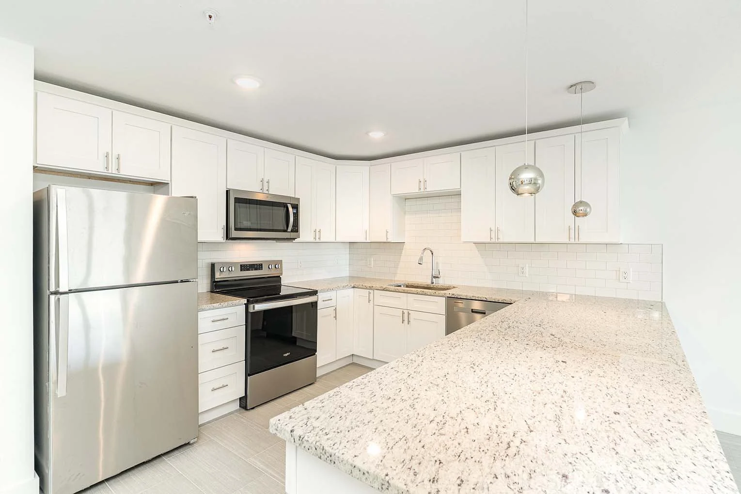 Modern kitchen with white cabinets, stainless steel refrigerator, microwave, oven, and dishwasher, beige granite countertops, white subway tile backsplash, sink with a faucet, and two hanging spherical pendant lights.
