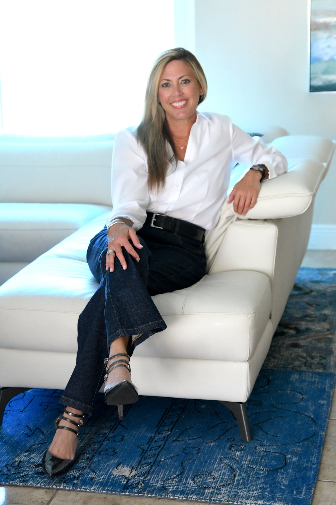 A woman with blonde hair, wearing a white blouse, black pants, and black heeled shoes, sitting on a white modern sofa in a well-lit room with a blue patterned rug and a painting on the wall.