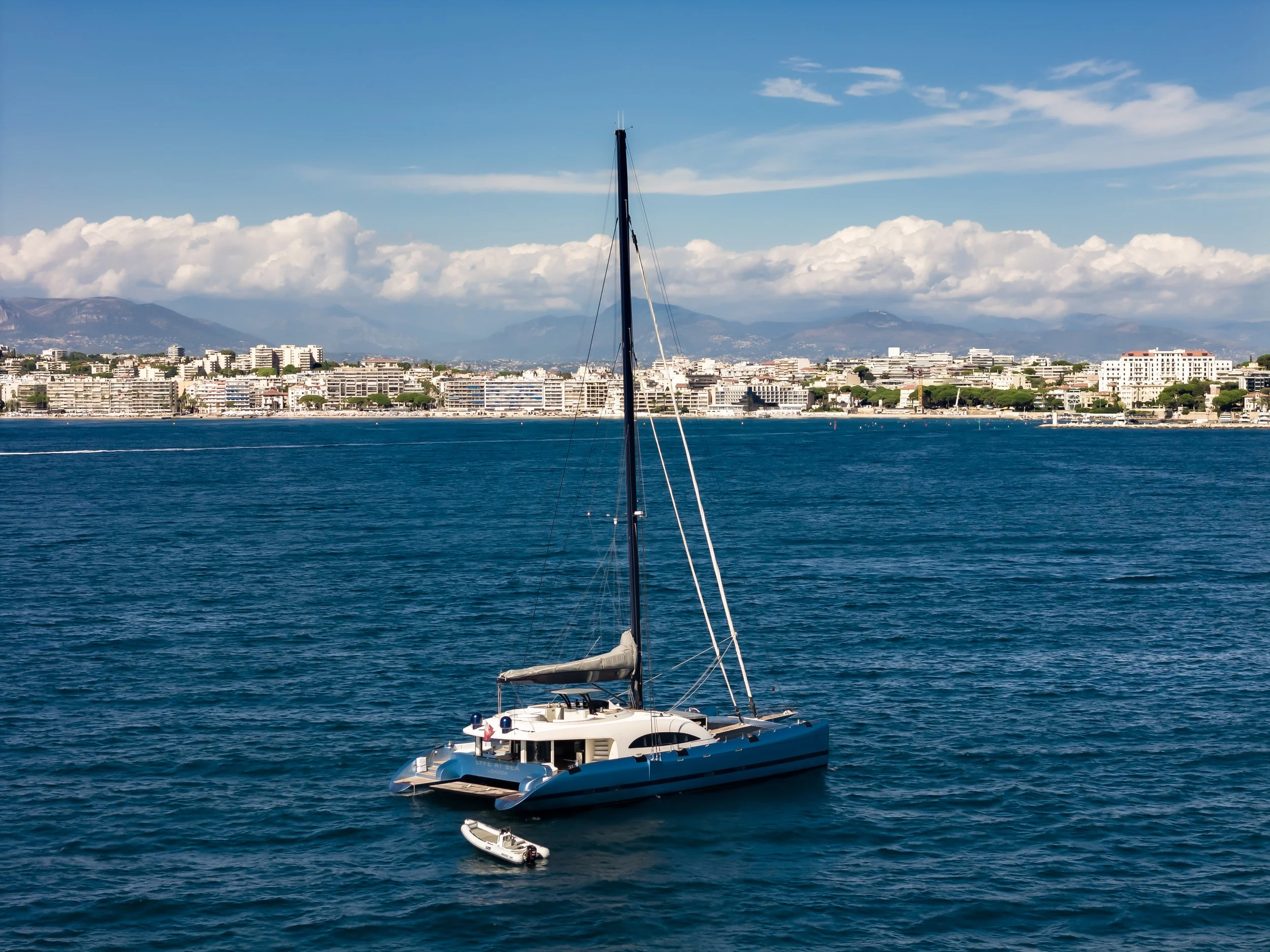 A large sailboat floating on calm ocean water with a cityscape and mountains in the background under a partly cloudy sky. LYFE AT SEA