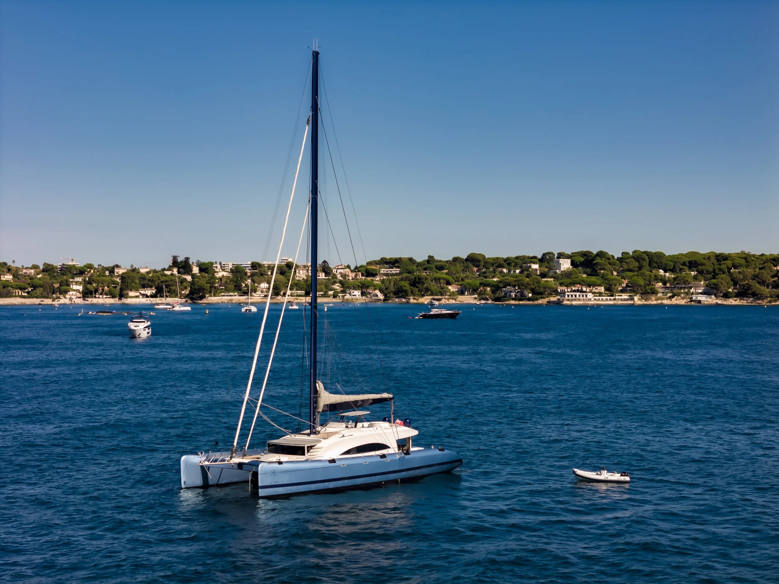 A white yacht sailing on deep blue water, with smaller boats nearby, and a green hillside with houses in the background under a clear blue sky. LYFE AT SEA
