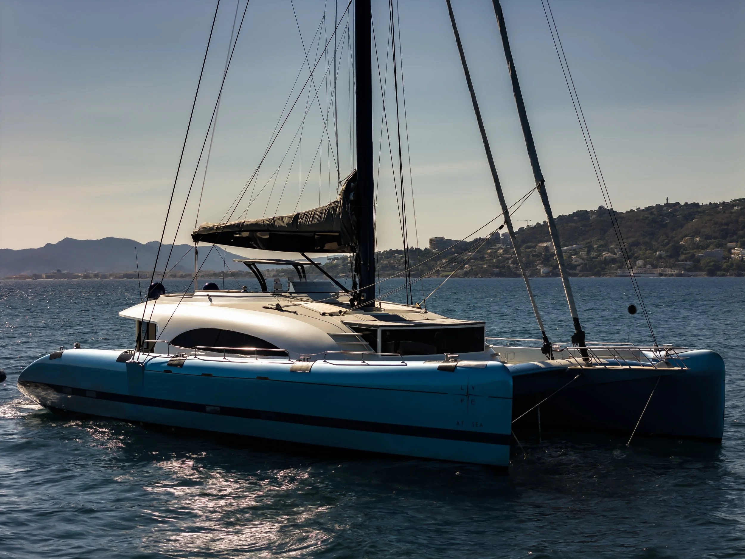 A sailboat with a black mast and white hull on calm water, with hills and buildings in the background during daytime. LYFE AT SEA