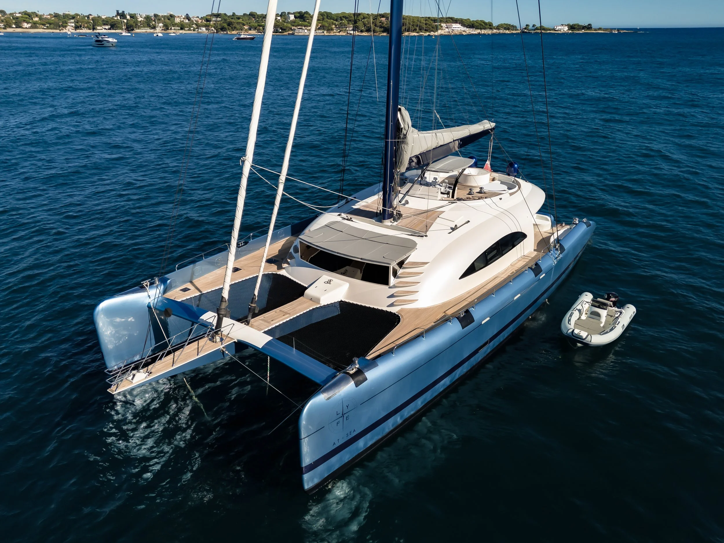 A luxurious white yacht sailing on calm blue water with several other boats in the distance and a lush green shoreline with houses and trees. LYFE AT SEA
