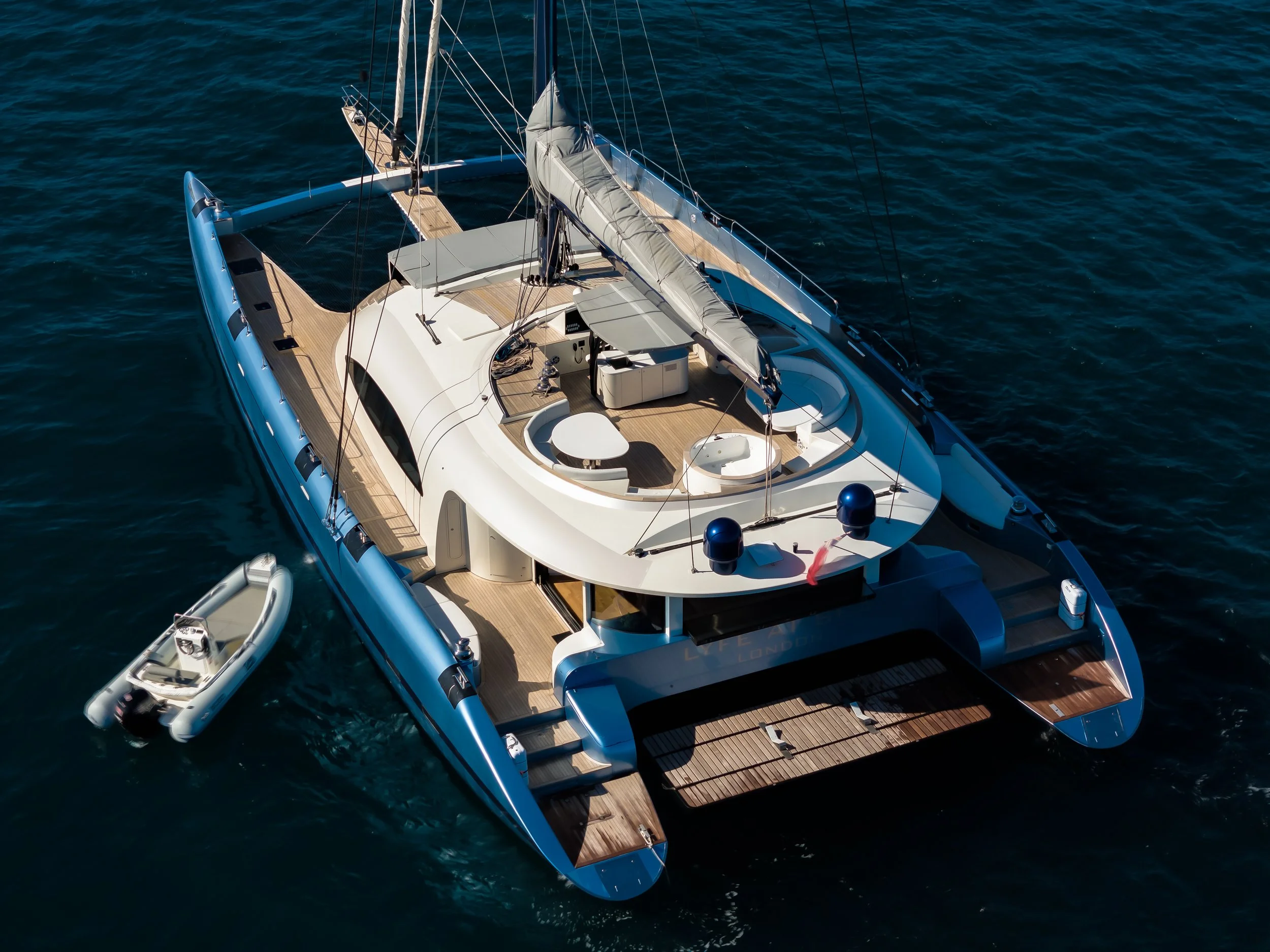 A luxury white sailing yacht with wooden accents floating on calm blue water, showing deck area, mast, and rigging from above. LYFE AT SEA