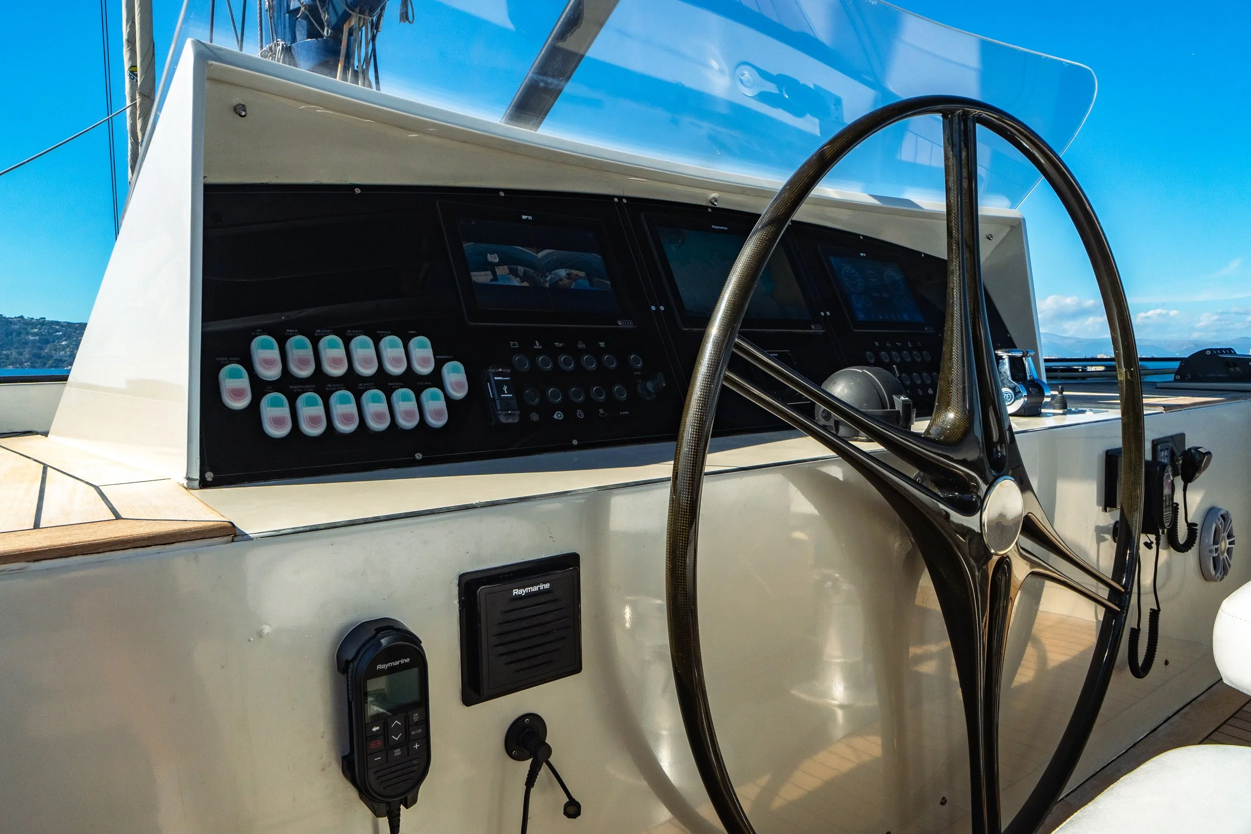Close-up of the dashboard and steering wheel of a yacht, with multiple digital displays, gauges, switches, and a radio control device, under a clear blue sky.