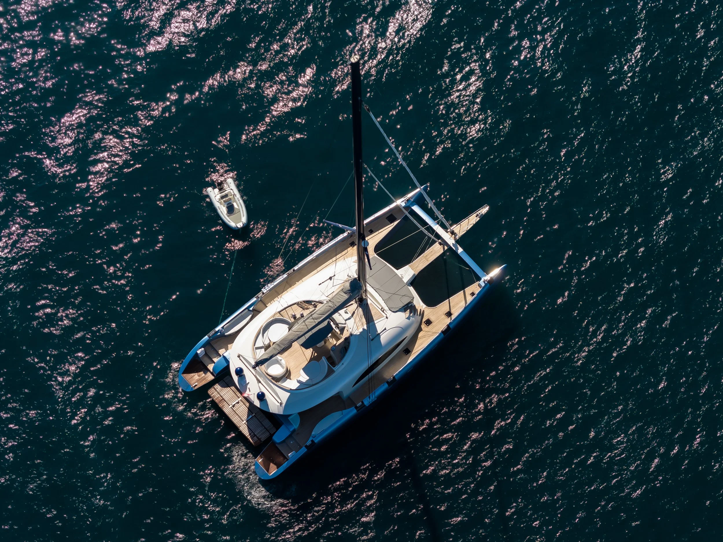 An aerial view of a sailboat with a smaller motorboat in the water, surrounded by dark blue ocean water with sunlight reflections. LYFE AT SEA