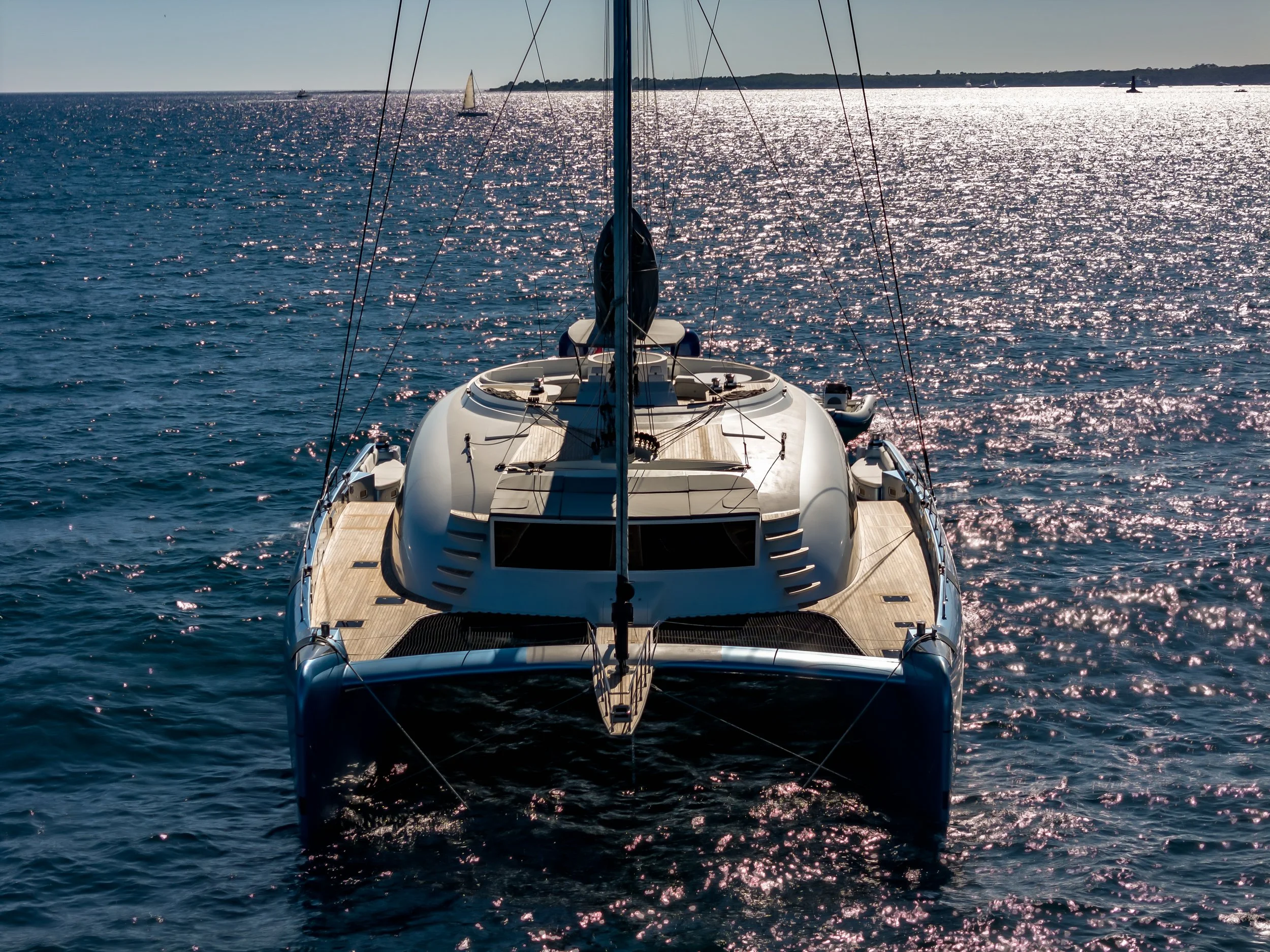 Aerial view of a luxury sailboat on calm water, with a distant coastline and several other boats and ships, under clear sky and bright sunlight.