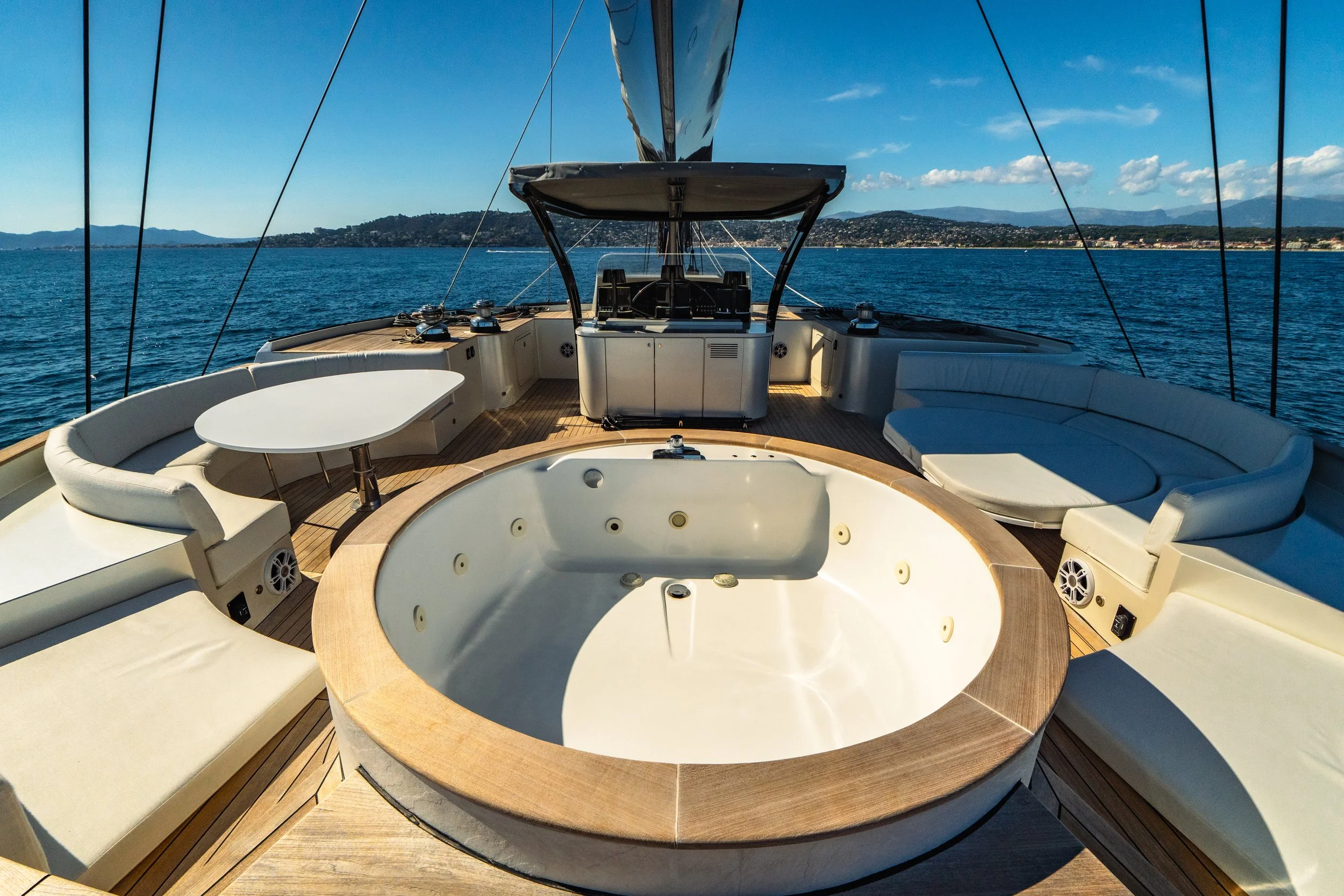 View from the yacht showing a large Jacuzzi surrounded by cream-colored cushioned seating on a wooden deck, with the ocean, distant shoreline, and blue sky with some clouds in the background. LYFE AT SEA