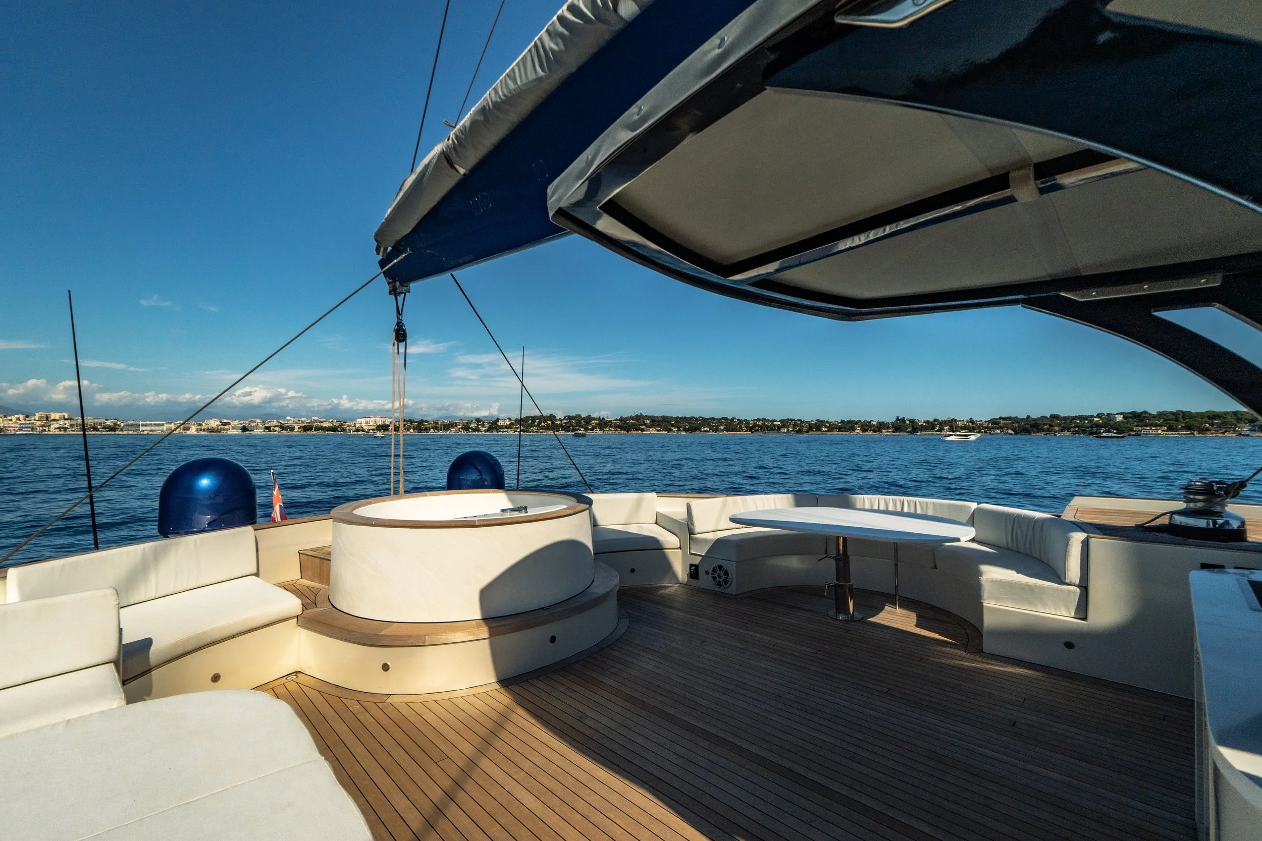 A view from a boat showing an outdoor lounge area with white cushioned seating, a round table, and a view of the water and city skyline in the distance under a clear blue sky.