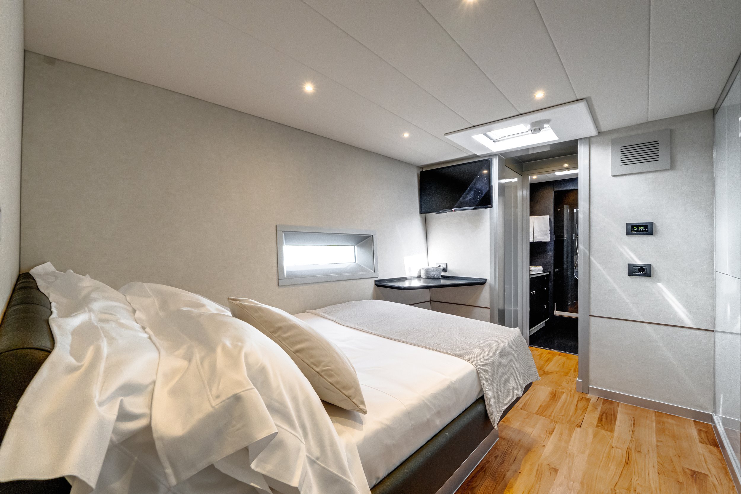A modern hotel room with a bed, a wall-mounted TV, a small window, and an adjacent bathroom with a glass shower door.