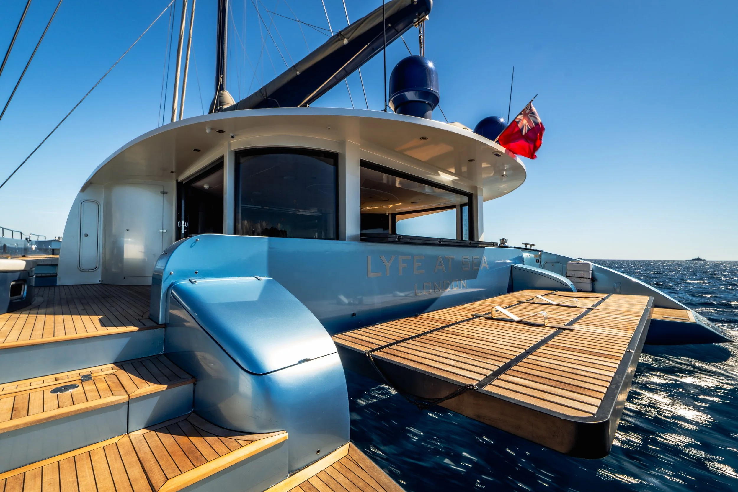 Close-up of a luxury yacht's deck, featuring wooden planks, sliding glass door, and a British flag, with the sea and blue sky in the background.