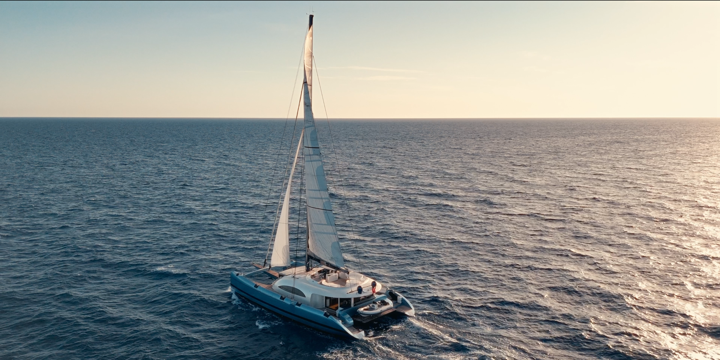A modern sailboat with white sails sailing on calm ocean water during sunset.