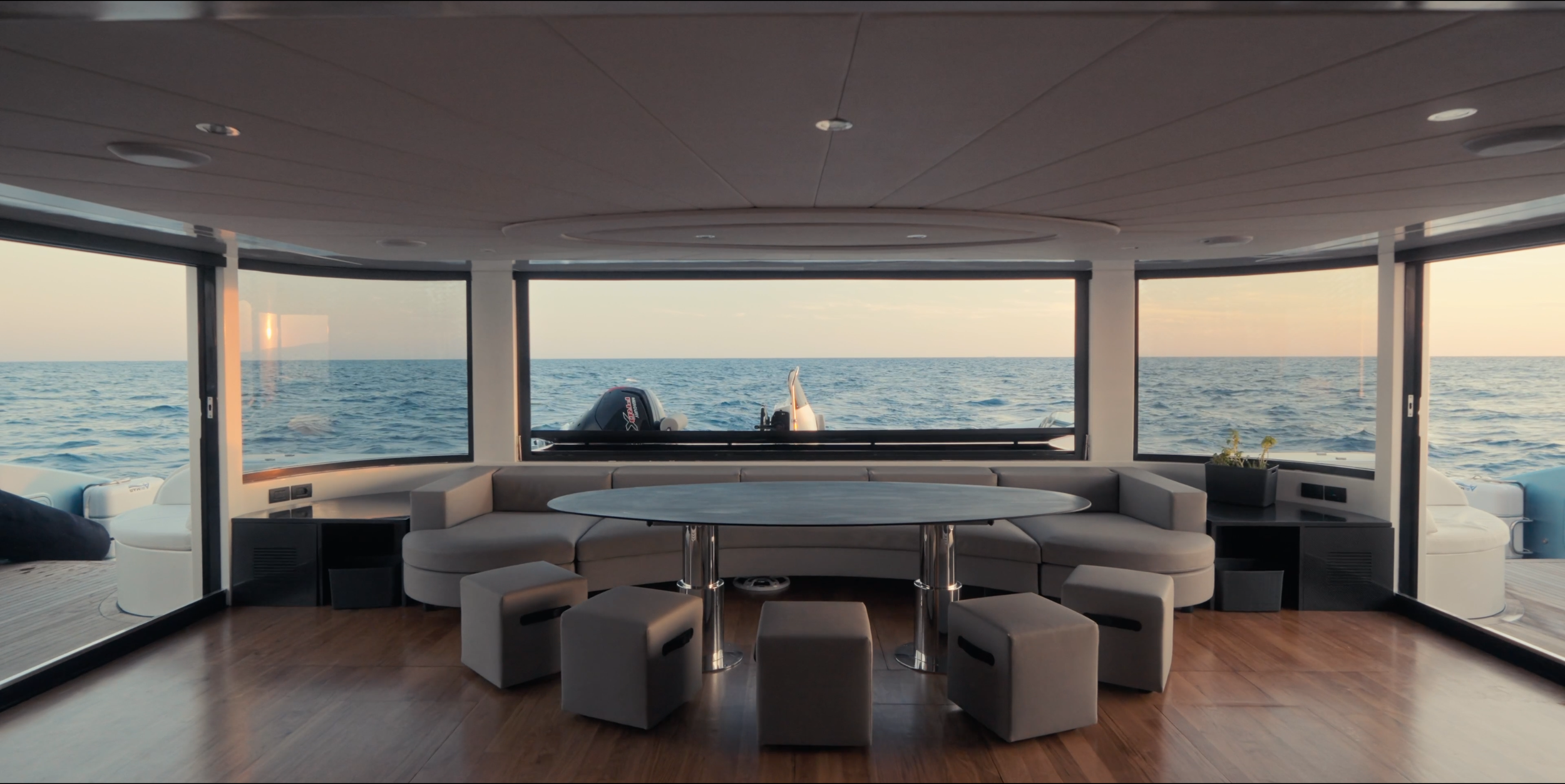 Interior of a yacht's living area with large windows showing the ocean and sunset, featuring a round table, cushioned seating, potted plant, and wood flooring.