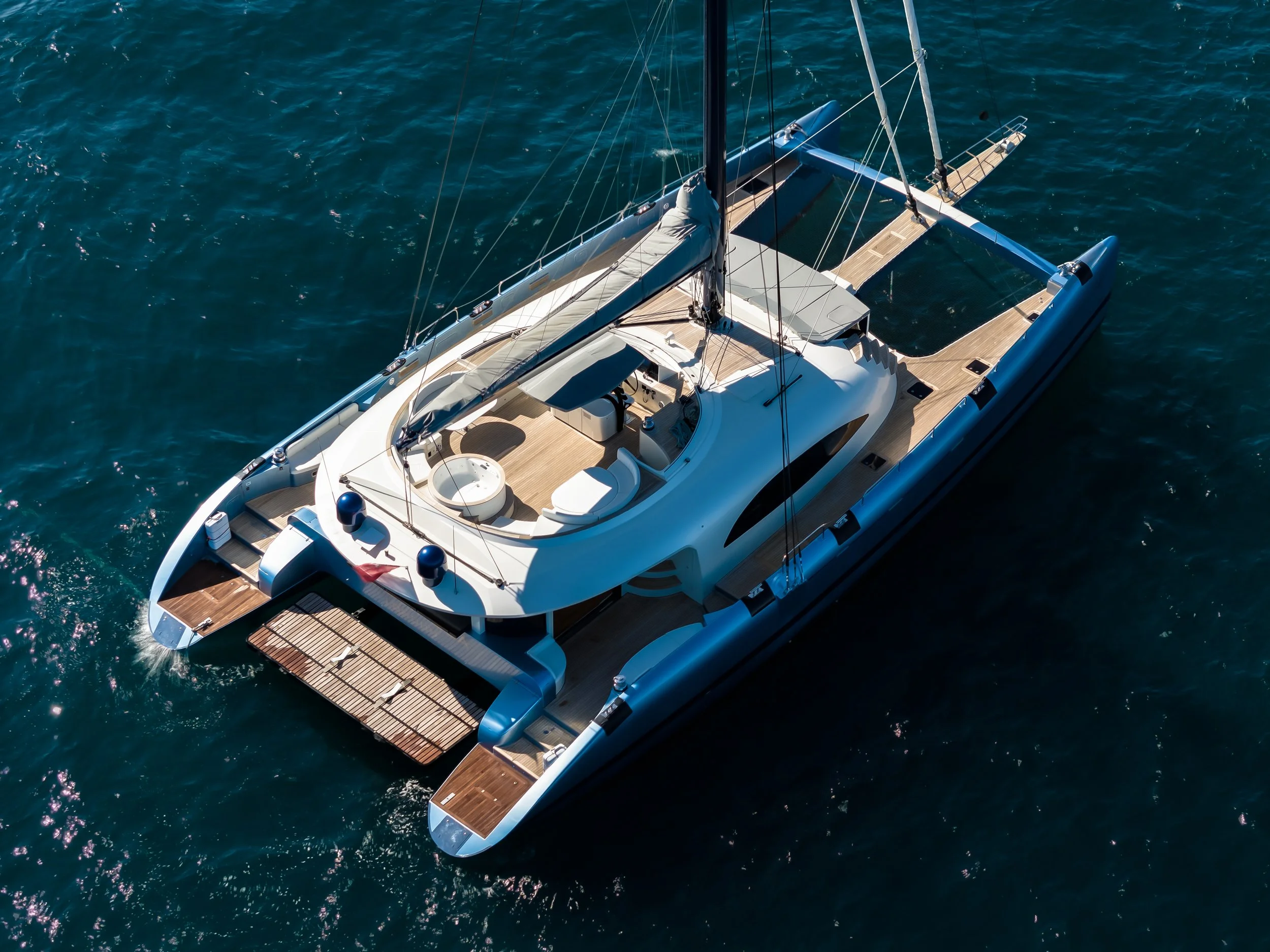 Overhead view of a luxurious sailboat on the ocean, featuring a spacious deck with seating areas, a covered helm station, and a large mast with sails. LYFE AT SEA