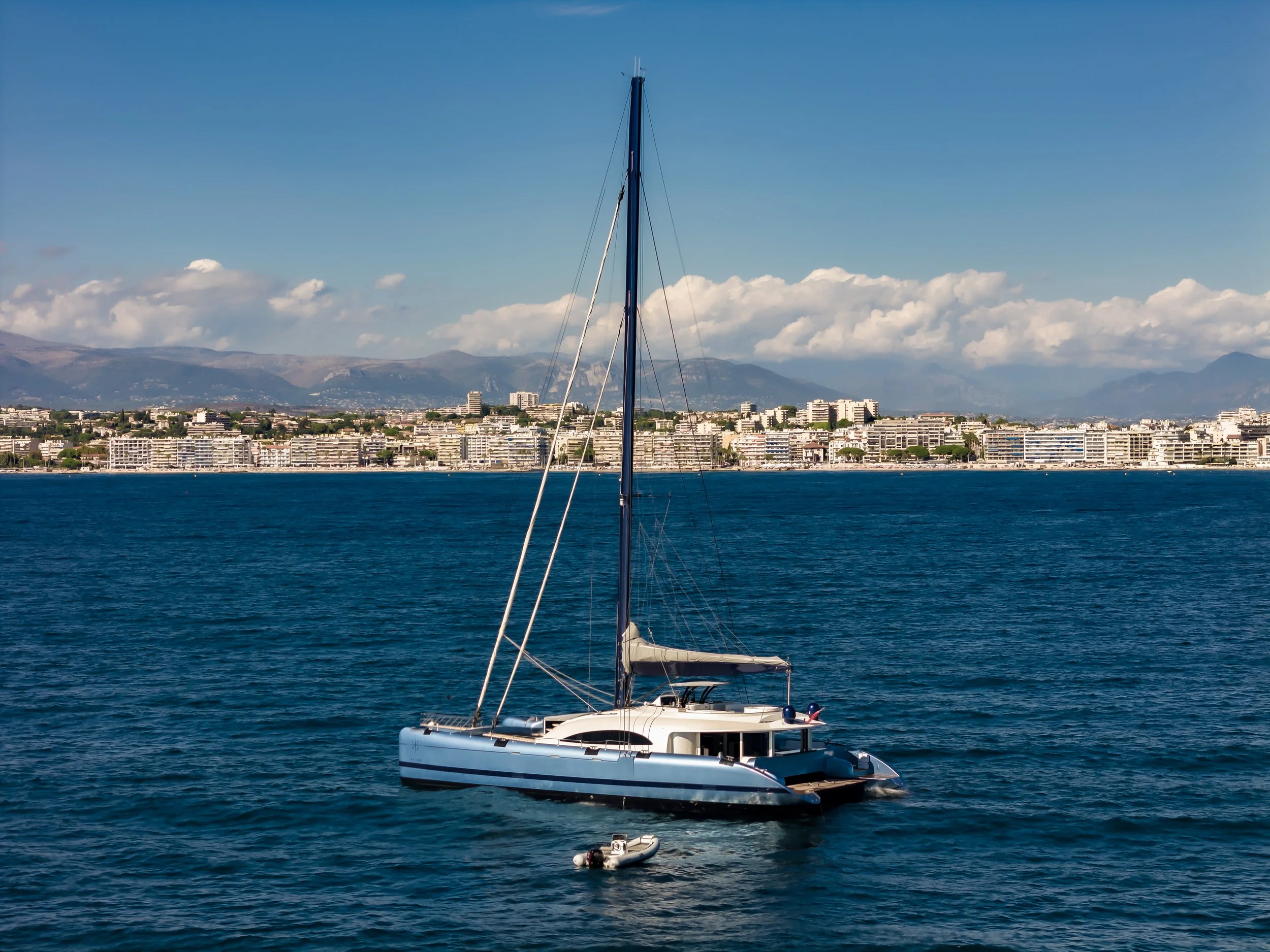 A white sailboat floating on the blue water with a cityscape and mountains in the background under a clear sky. LYFE AT SEA