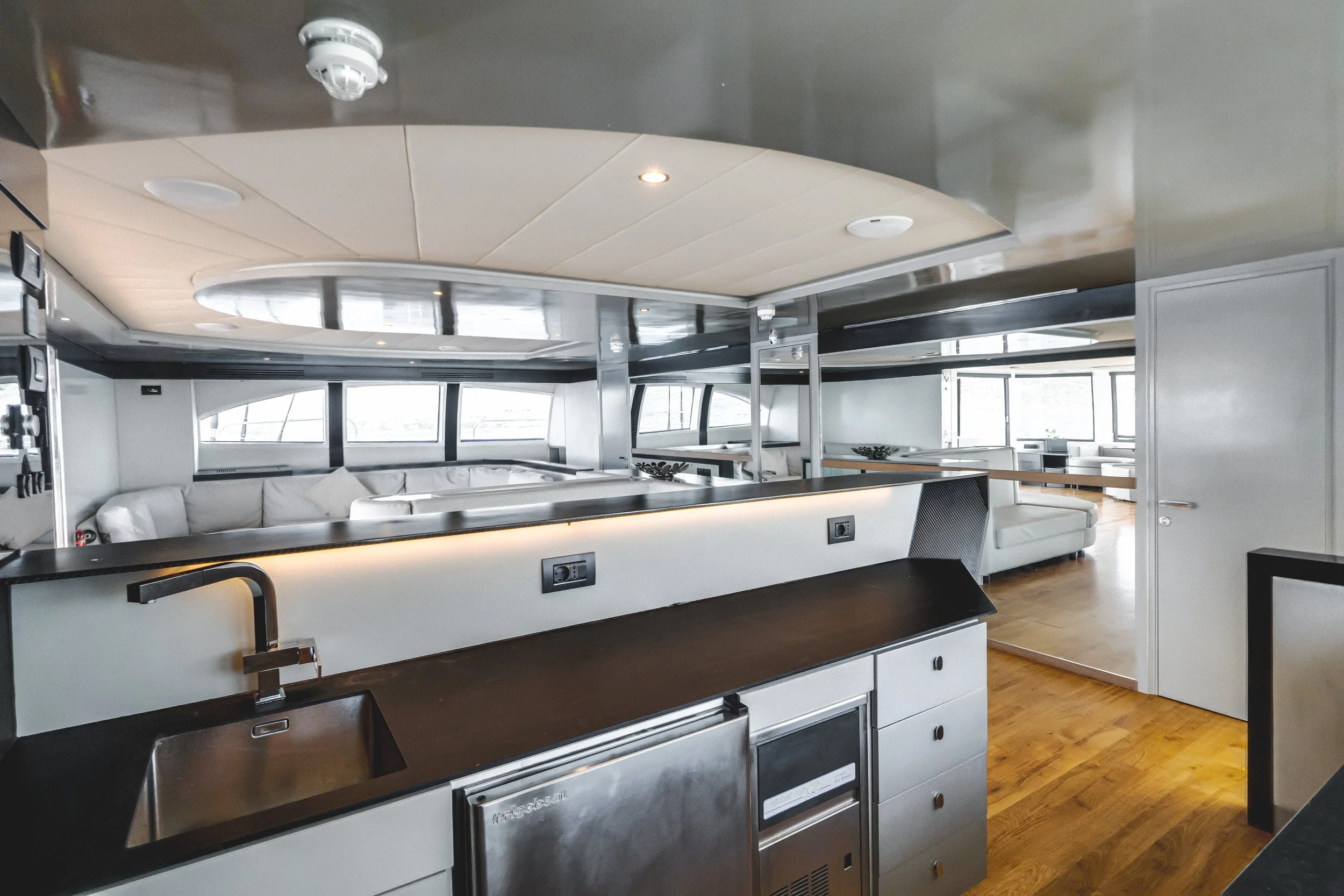Modern interior of a yacht or boat with a curved ceiling, large windows, white seating area, black and white kitchen counter with appliances, and wooden flooring. LYFE AT SEA