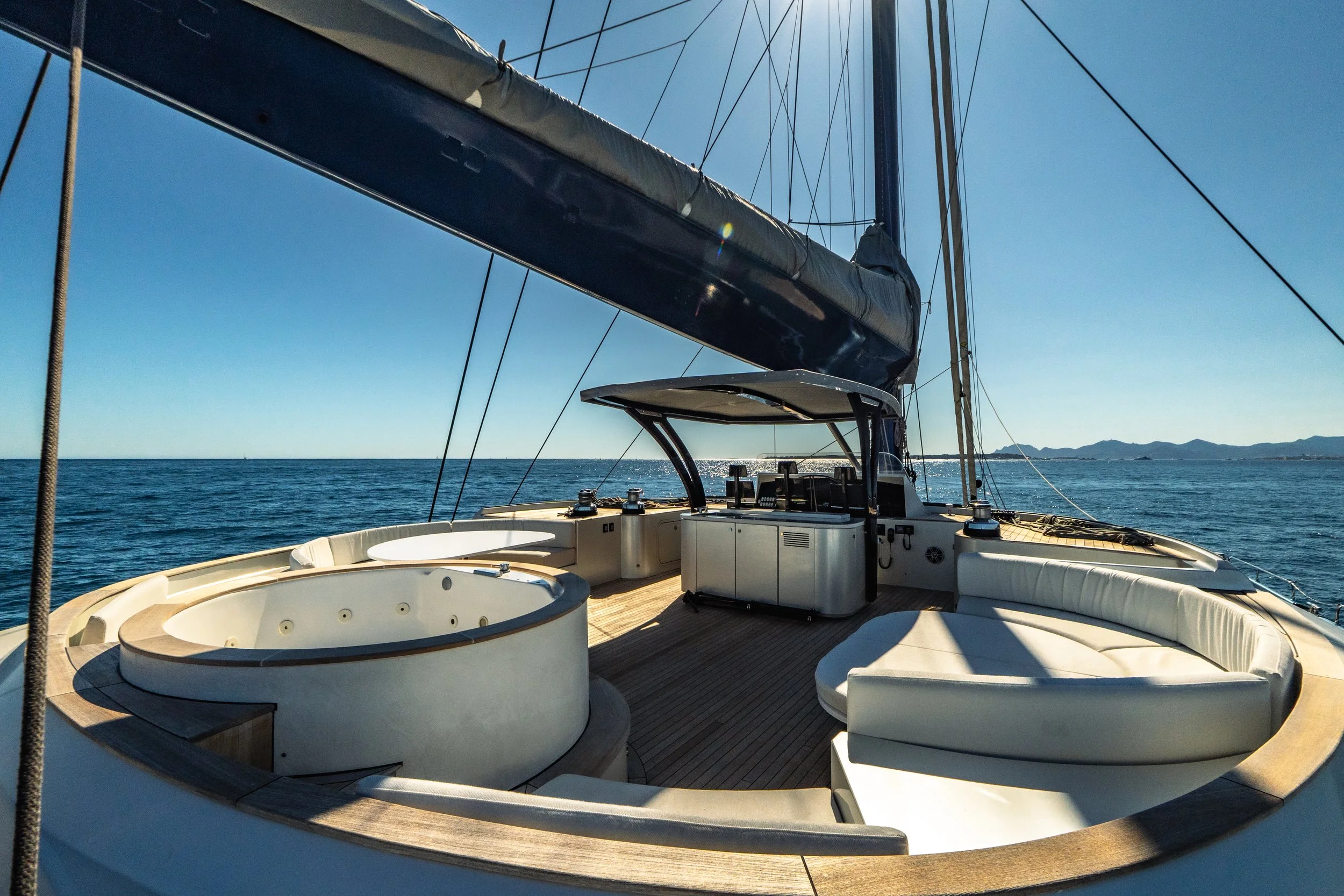 View from the deck of a luxury yacht showing a hot tub, seating area, and a helm station with navigational equipment, with the ocean and distant islands in the background under a clear blue sky. LYFE AT SEA