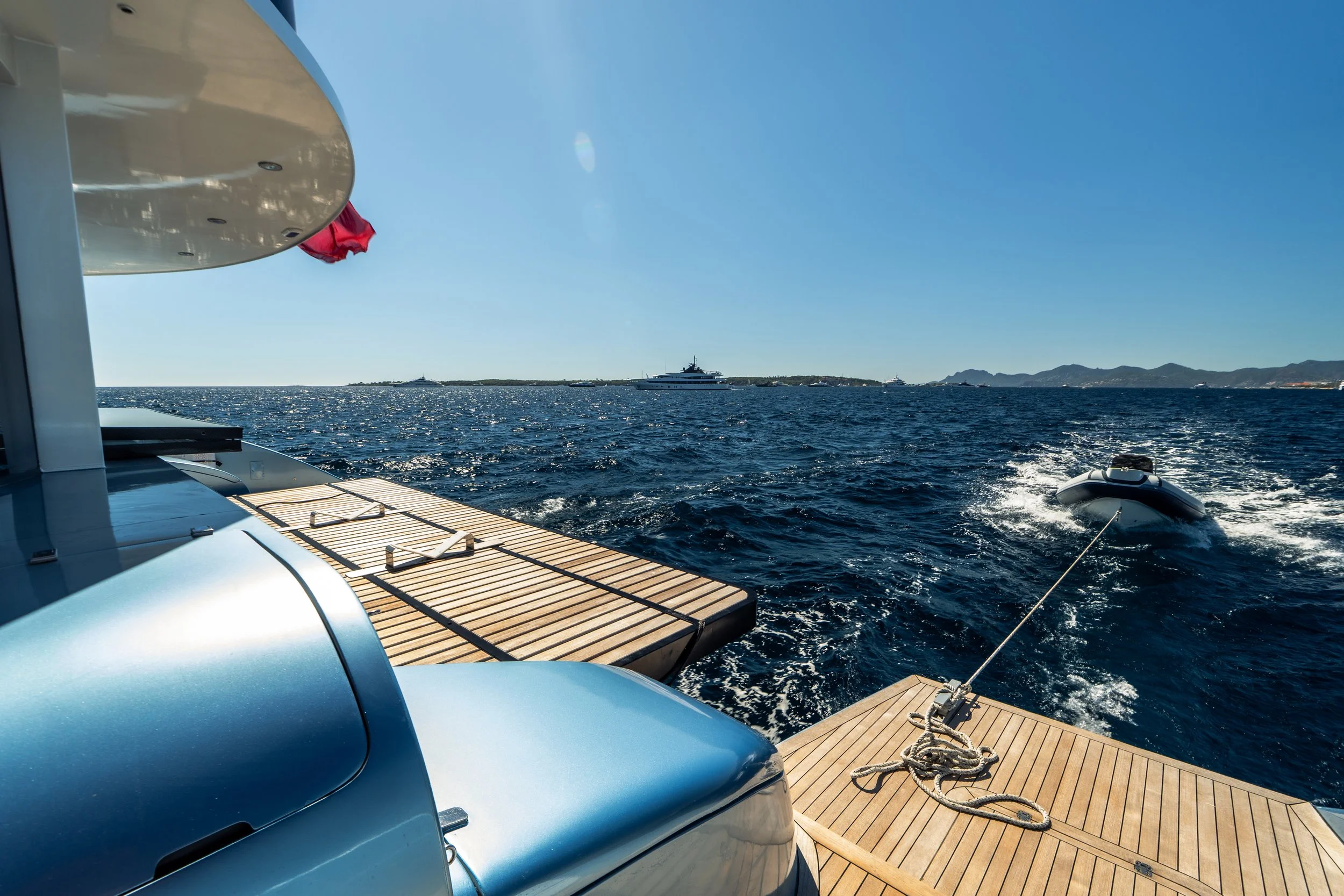 View from a yacht showing a wooden deck, a portion of the yacht's body, water, other yachts, and distant land with mountains under a clear blue sky.