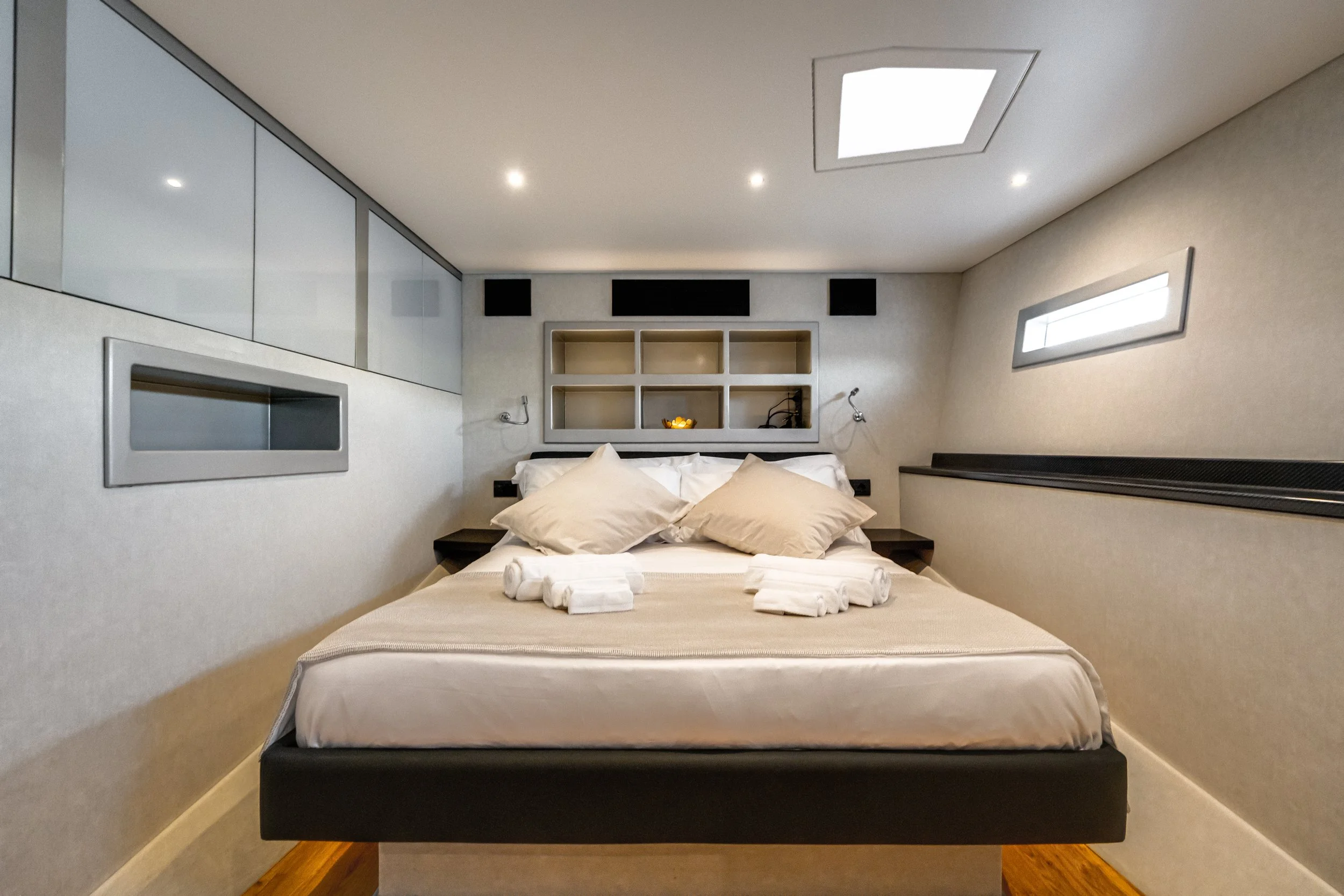 Modern bedroom with a bed, pillows, bedside table, built-in shelving behind bed, and ceiling skylight.