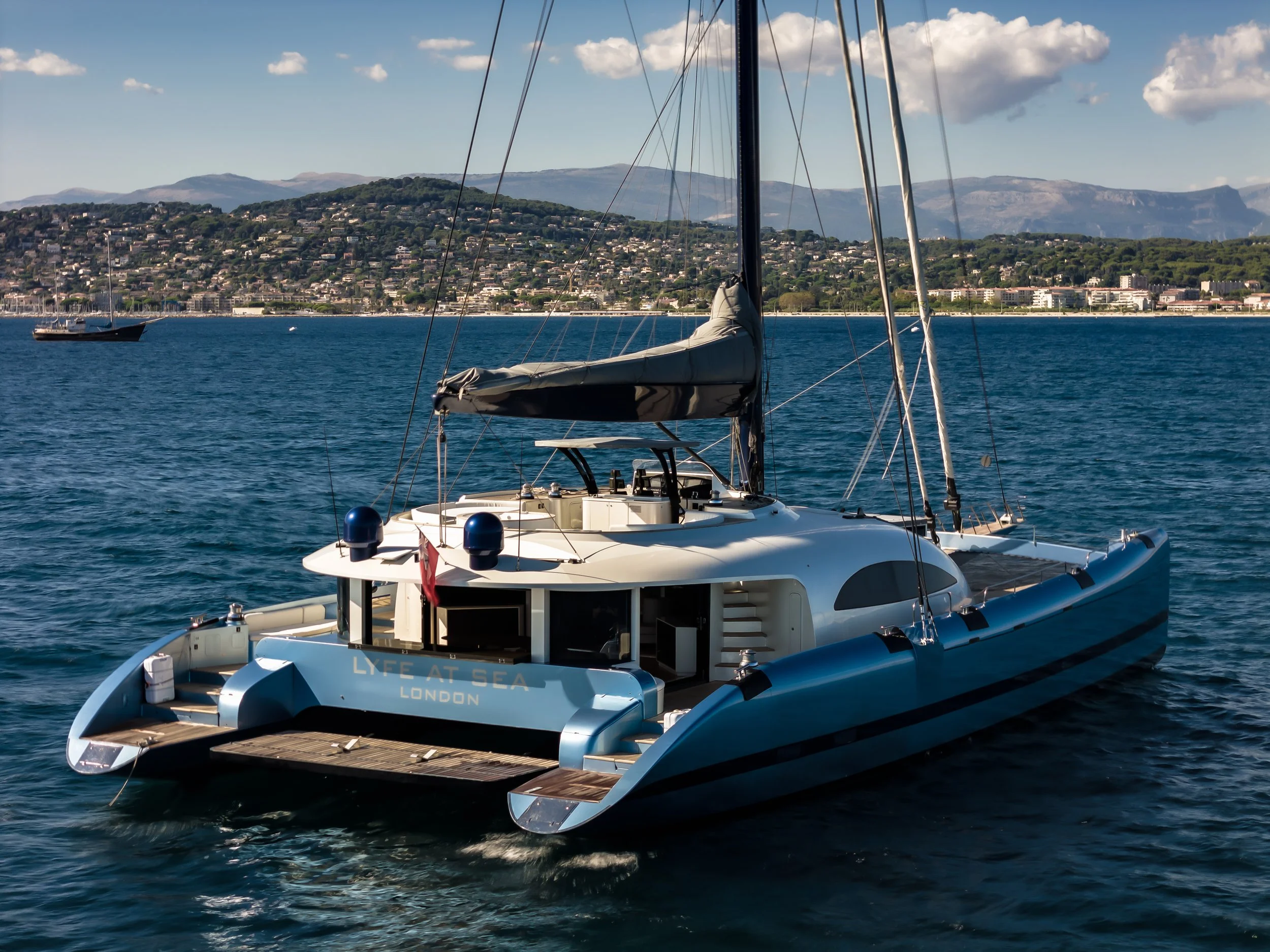 A luxury yacht named 'LYFE AT SEA' docked on calm water with a hilly shoreline and buildings in the background under a partly cloudy sky. LYFE AT SEA