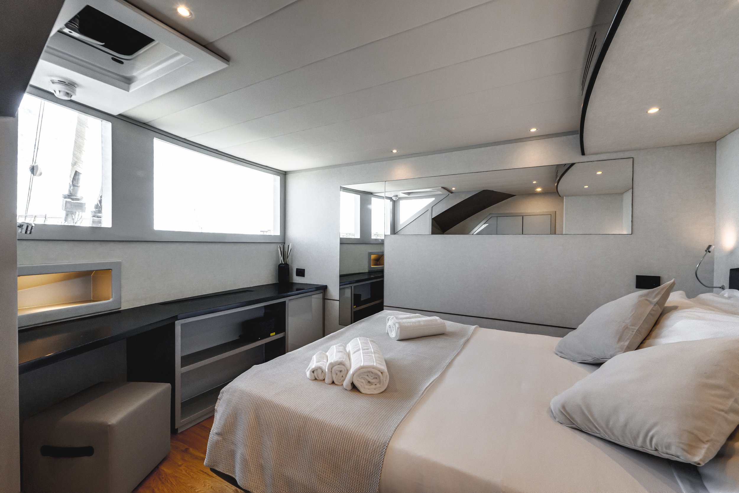 A modern hotel room featuring a large bed with white linens and rolled towels, a built-in black counter with storage, large windows with natural light, and a mirror on the wall reflecting some of the room's ceiling and lighting. LYFE AT SEA