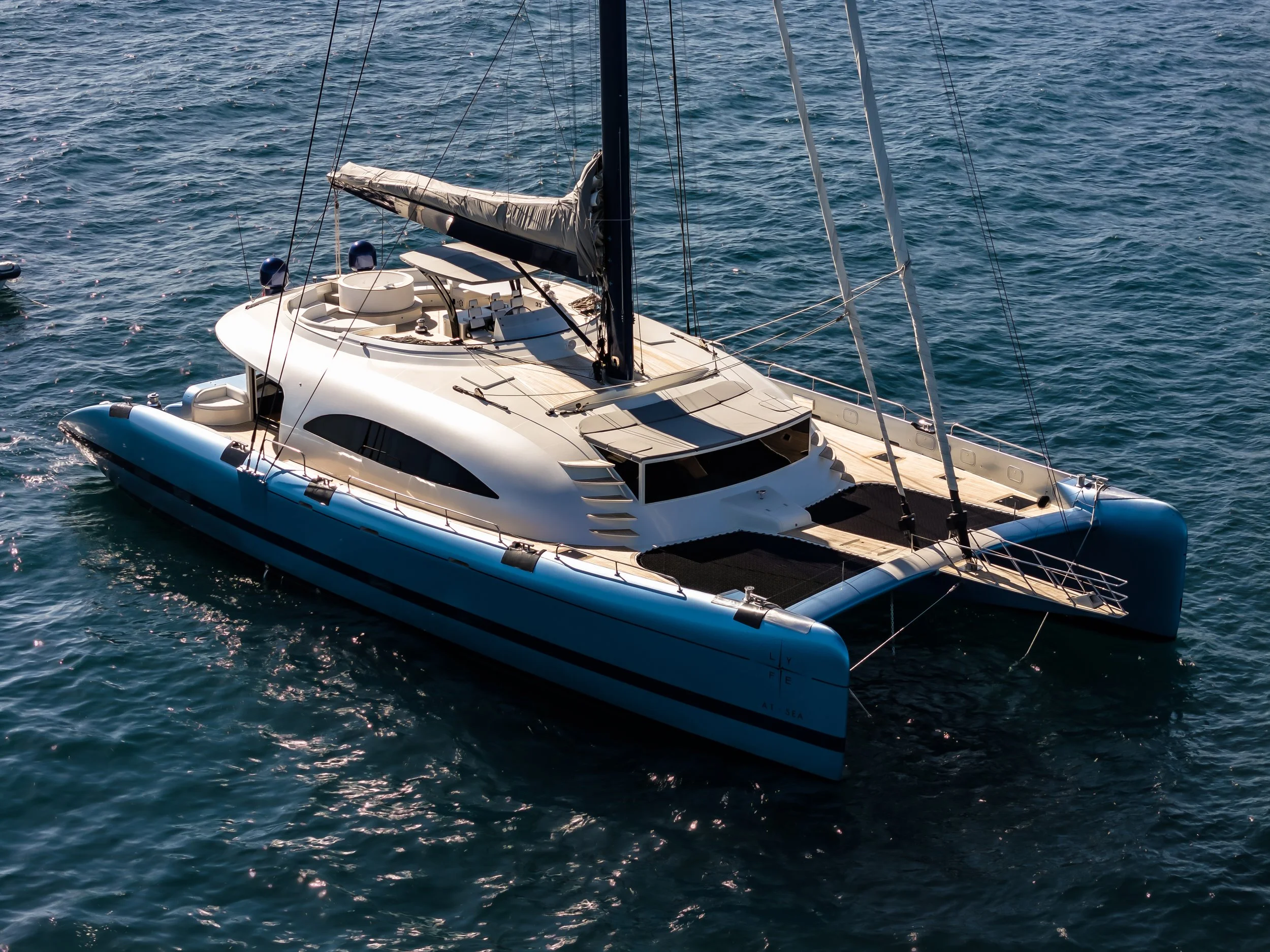A luxury white sailboat with a black mast and beige deck, floating on calm blue ocean water. LYFE AT SEA