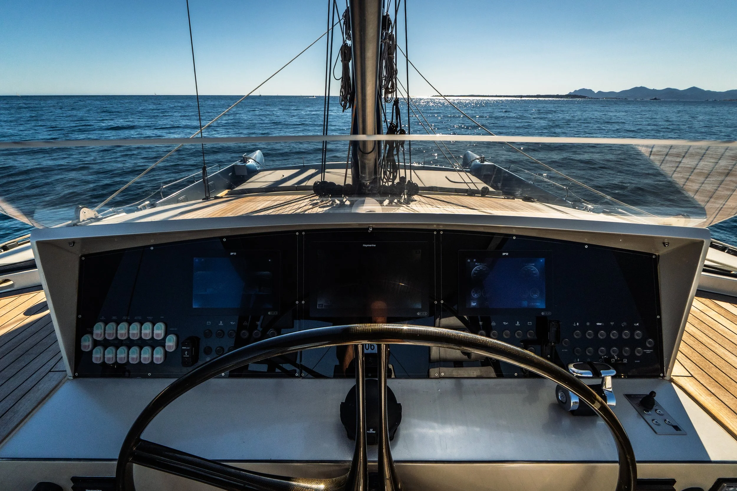 Inside view of a modern sailing yacht's cockpit with navigation display screens and control panels, looking out over the ocean with a distant coastline and hills under a clear sky.