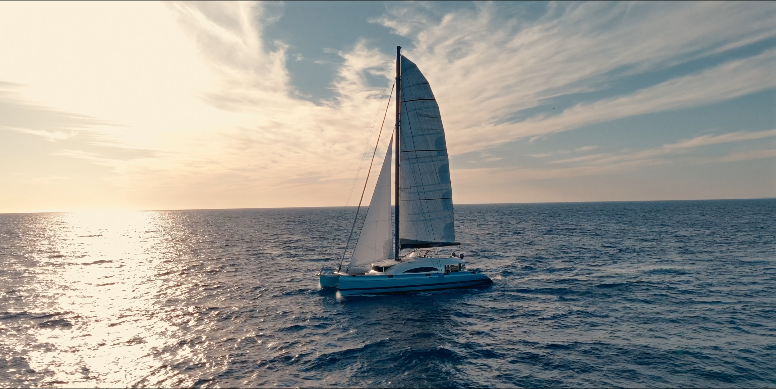 A sailboat with white sails and a blue spinnaker sailing on the ocean during sunset with a partly cloudy sky. LYFE AT SEA