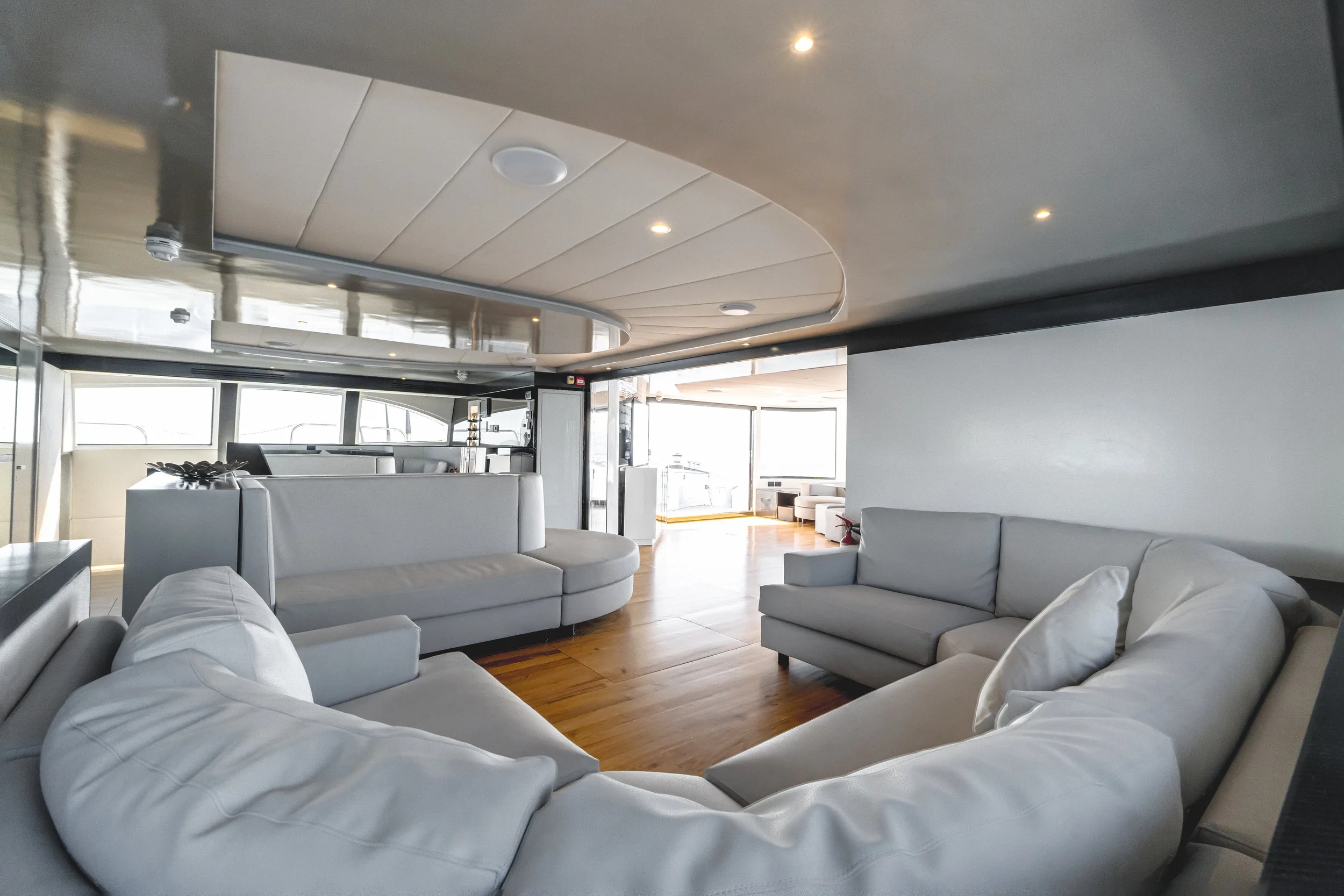 A spacious modern living room with white sofas arranged in a semi-circle, wooden flooring, large windows letting in natural light, and a curved ceiling with recessed lights. LYFE AT SEA