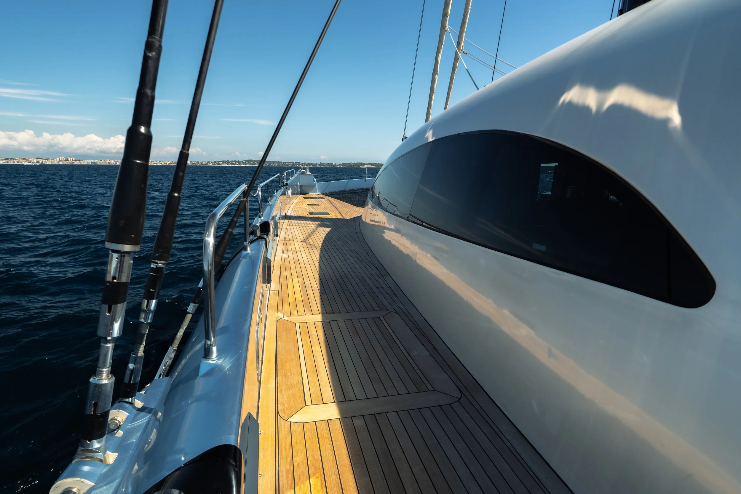 Side deck of a modern yacht with fishing rods, polished white hull, wooden flooring, and ocean water, with a distant city skyline and blue sky in the background. LYFE AT SEA