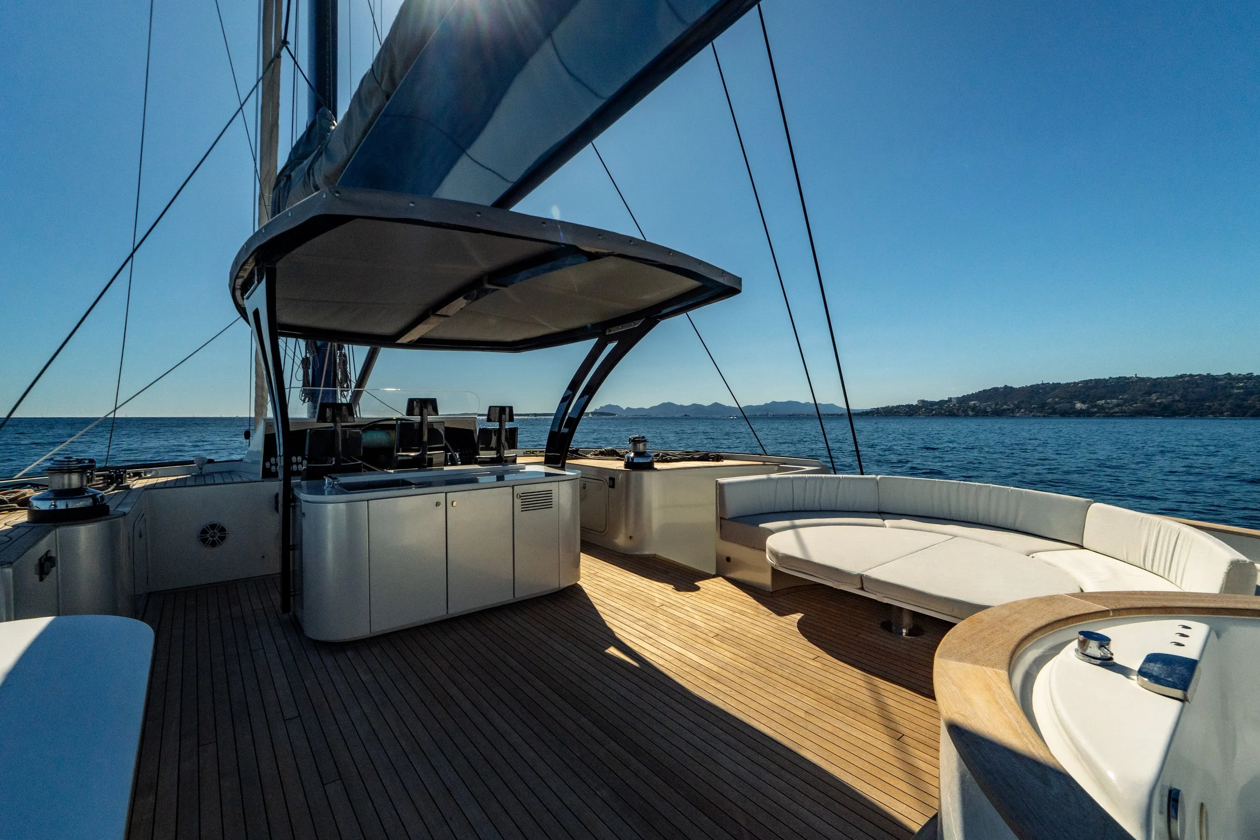 View from the deck of a yacht or boat, showing seating area with white cushioned seats, a small round table, and navigation equipment. The background features calm blue waters, distant land, and clear blue sky with sunlight. LYFE AT SEA