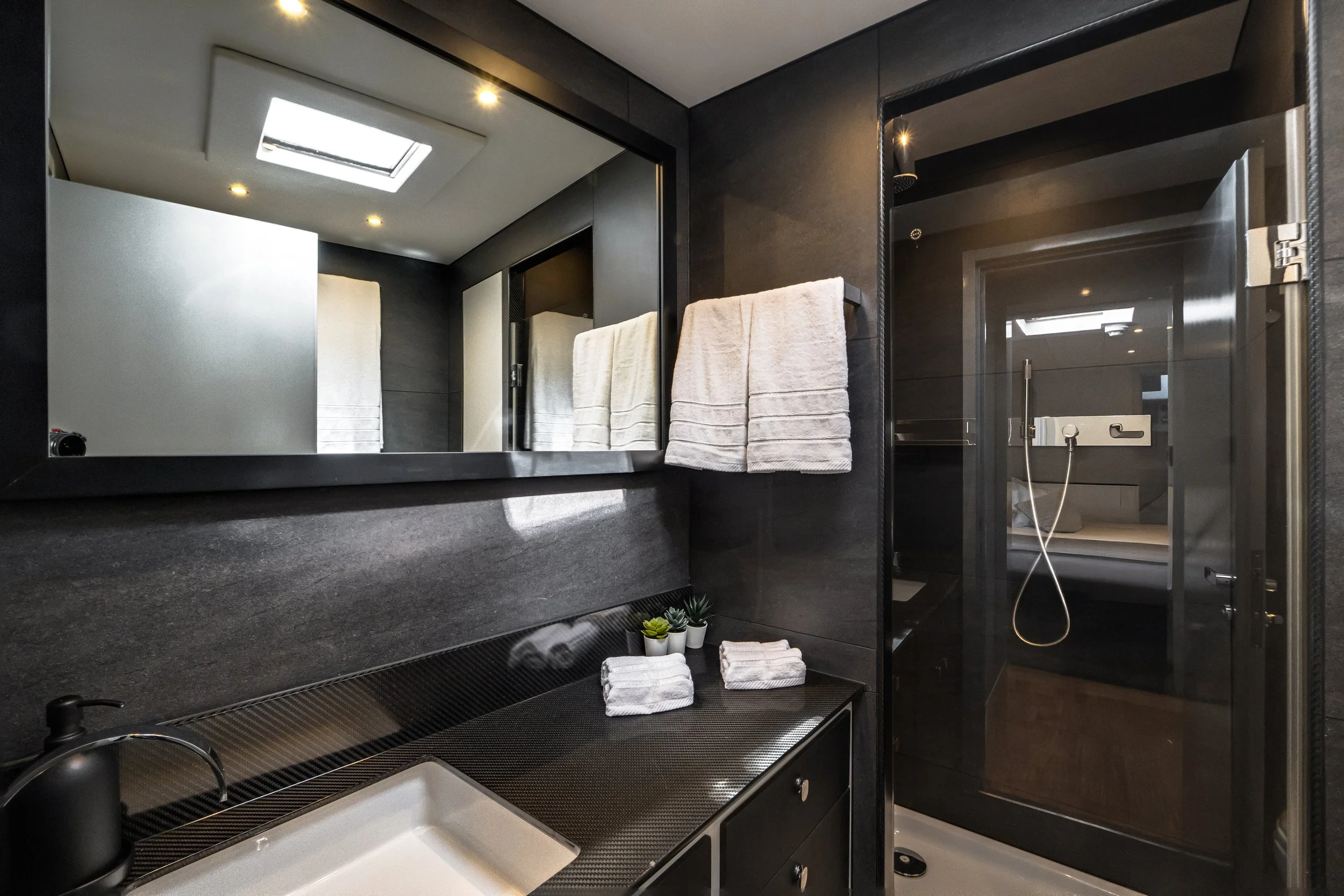 Modern bathroom with black and white color scheme, including a large mirror, black countertop with a sink, folded white towels, small potted plants, and a shower area with a glass door and a skylight on the ceiling.