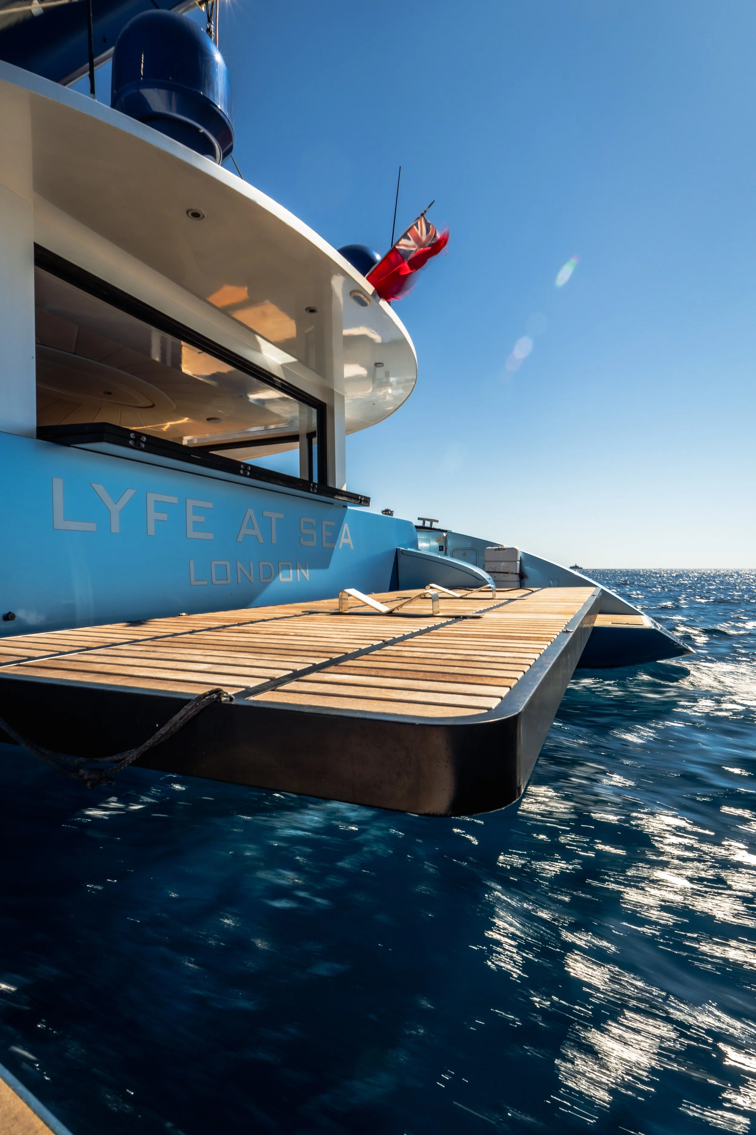 Close-up view of the deck of a luxury yacht named 'LYFE AT SEA', with part of the yacht's white structure and a small red flag, in the ocean on a sunny day with clear blue sky.