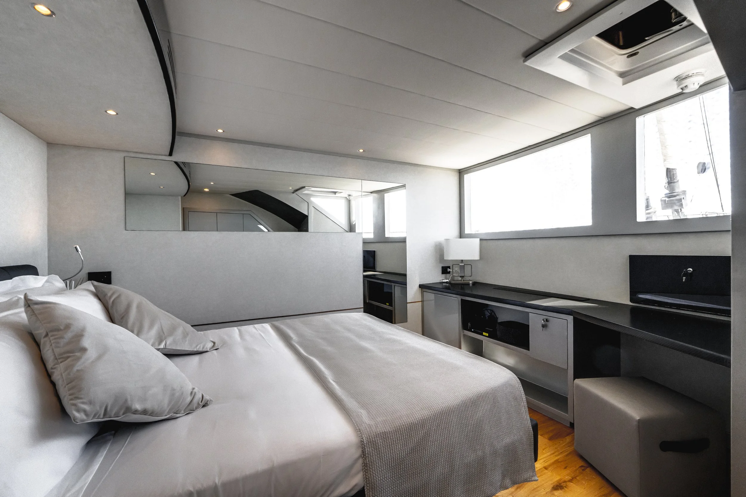 A modern hotel room with a white bed and multiple pillows, large windows, and minimalist furniture including a desk with a lamp and a monitor. LYFE AT SEA