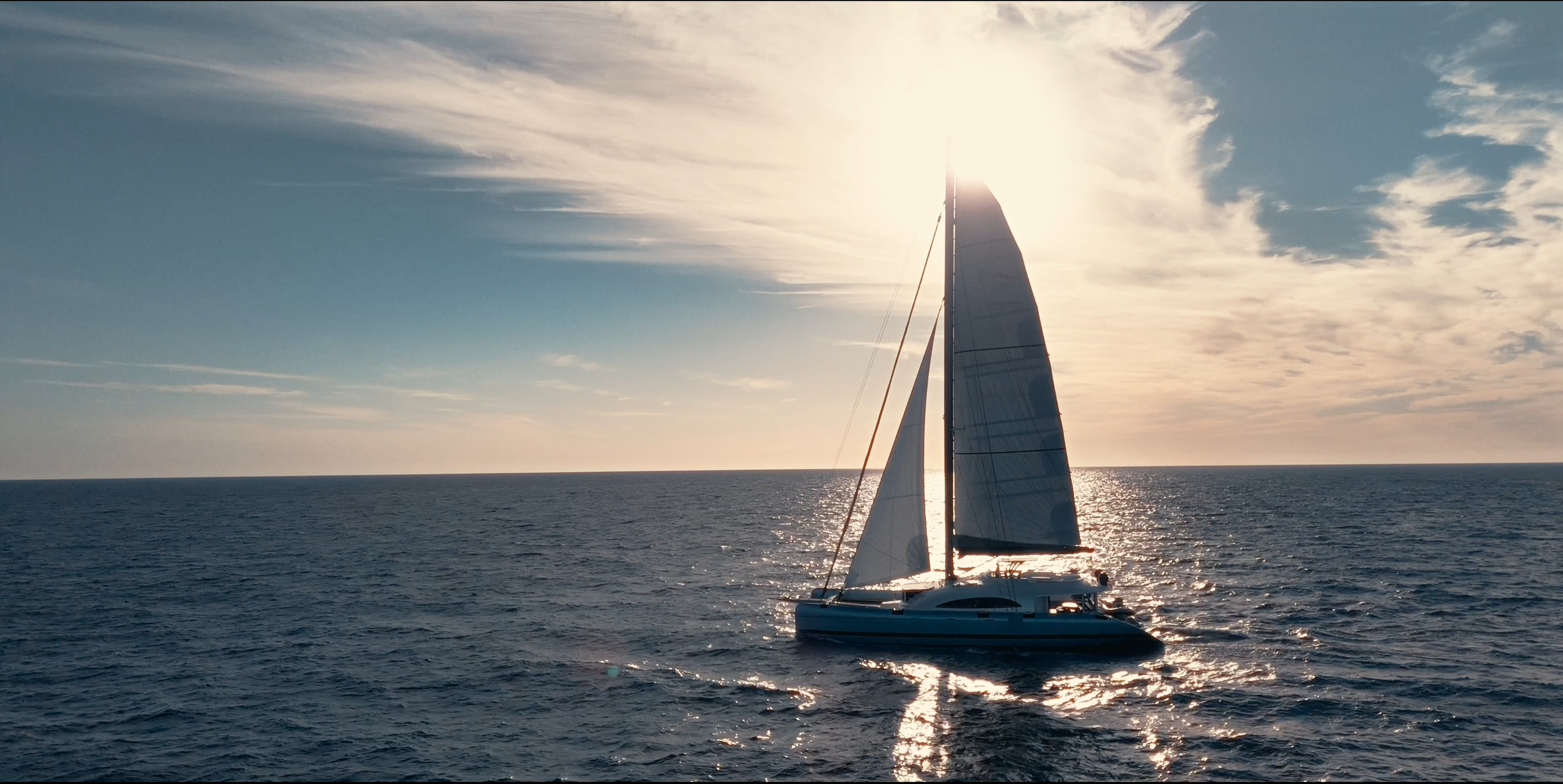 A sailboat on the ocean during sunset with the sun partially hidden behind the sail, creating a reflection on the water and a partly cloudy sky. LYFE AT SEA