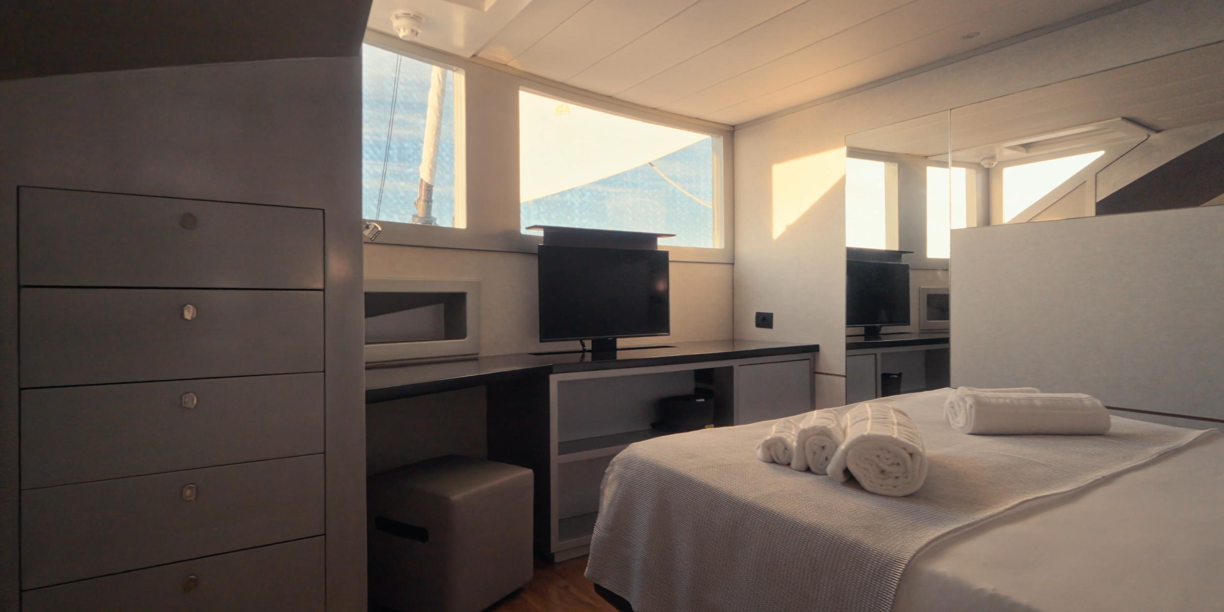 Interior of a modern hotel room with a bed, rolled towels, a desk with a small TV, and windows letting in natural light. LYFE AT SEA