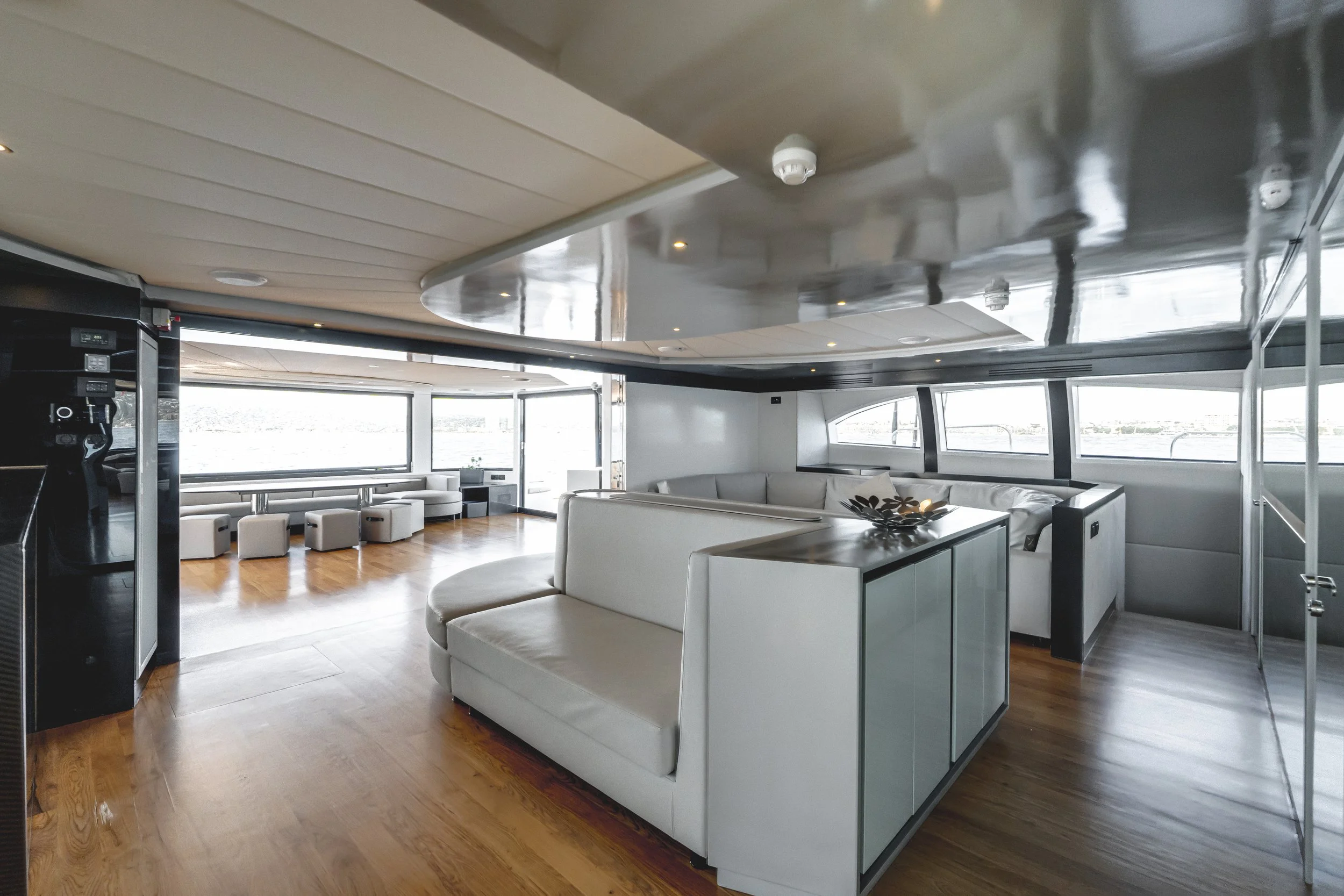 Interior of a yacht with white curved sofas, side tables, and large windows showing water outside. Ceiling has built-in lights and smoke detectors. LYFE AT SEA