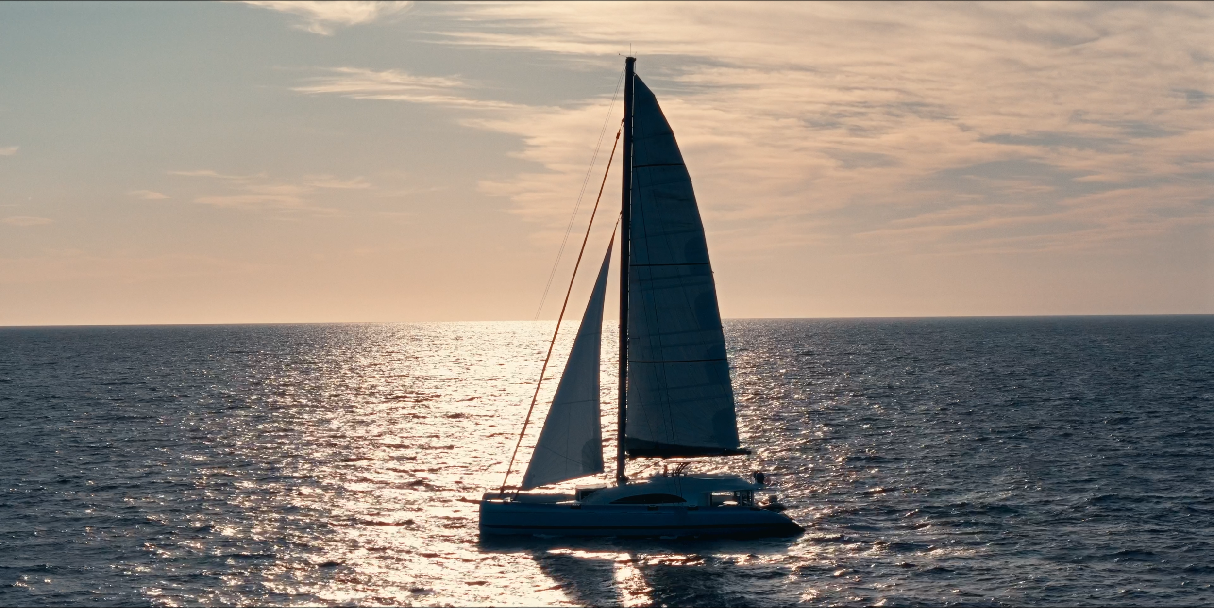 A sailboat with dark sails on the ocean during sunset, with the sky and water reflecting warm light.
