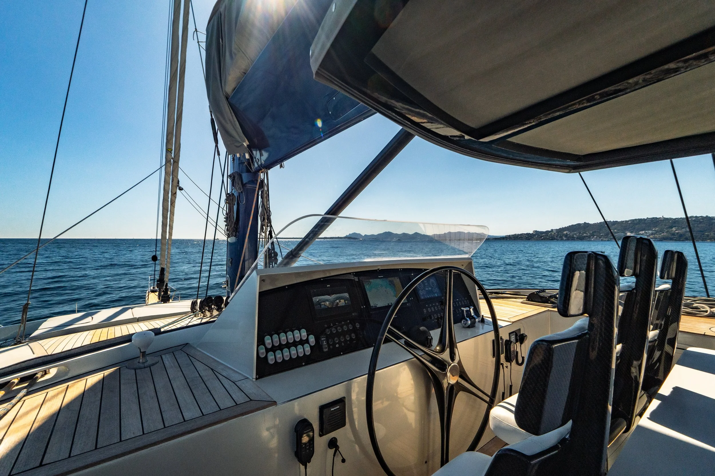 Interior view of a boat's cockpit with steering wheel and control panel, overlooking the ocean with a distant shoreline and islands under a clear blue sky. LYFE AT SEA