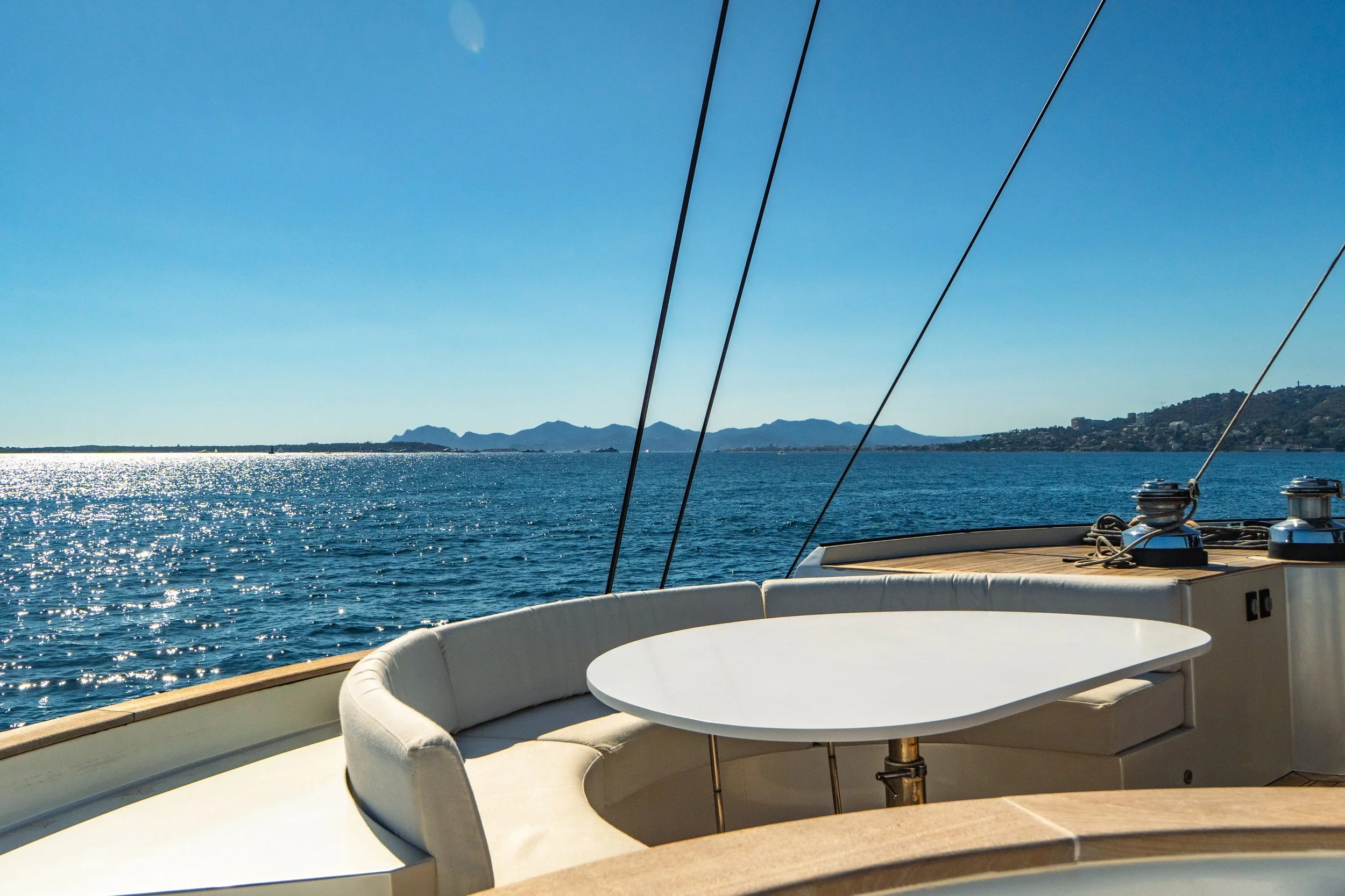 View from a boat showing the ocean, distant mountains, and clear blue sky with the moon visible. Part of the boat's rail, a white cushioned seating area, and a round white table are in the foreground.